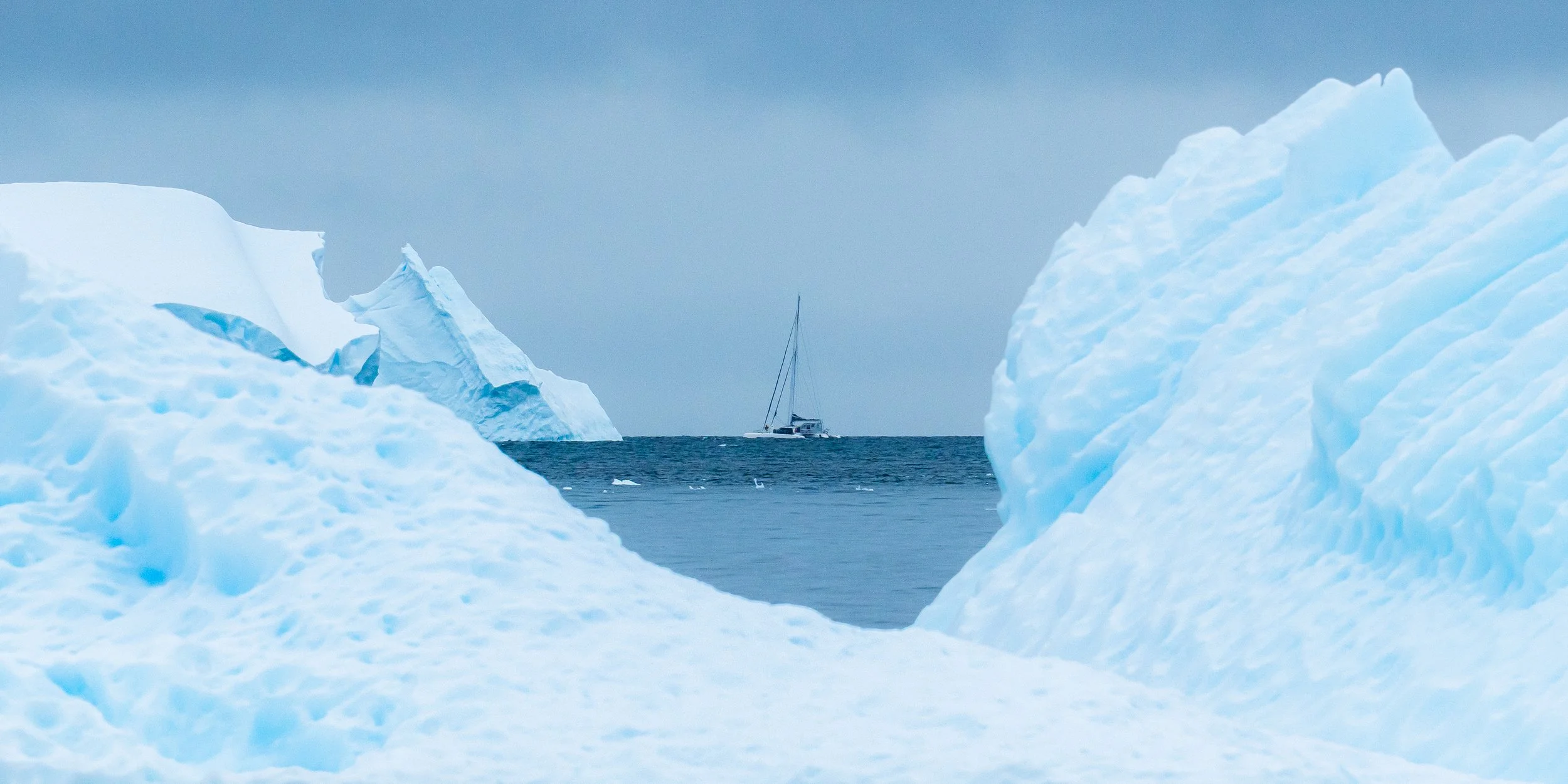 A sailboat on calm water seen between large icebergs in an icy seascape.