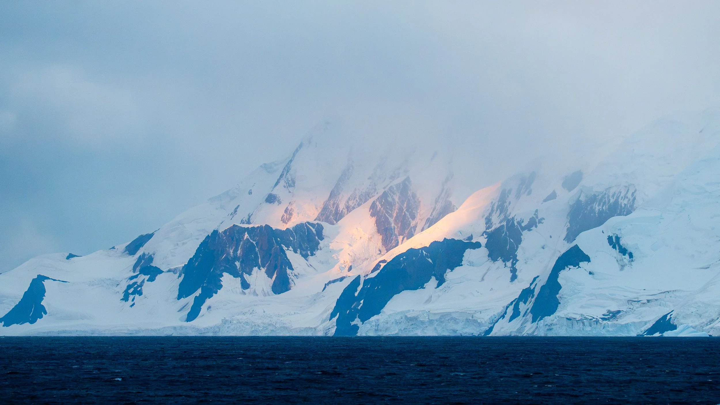 Snow-covered mountain range with sharp peaks, some illuminated by the setting or rising sun, and a body of water in the foreground.