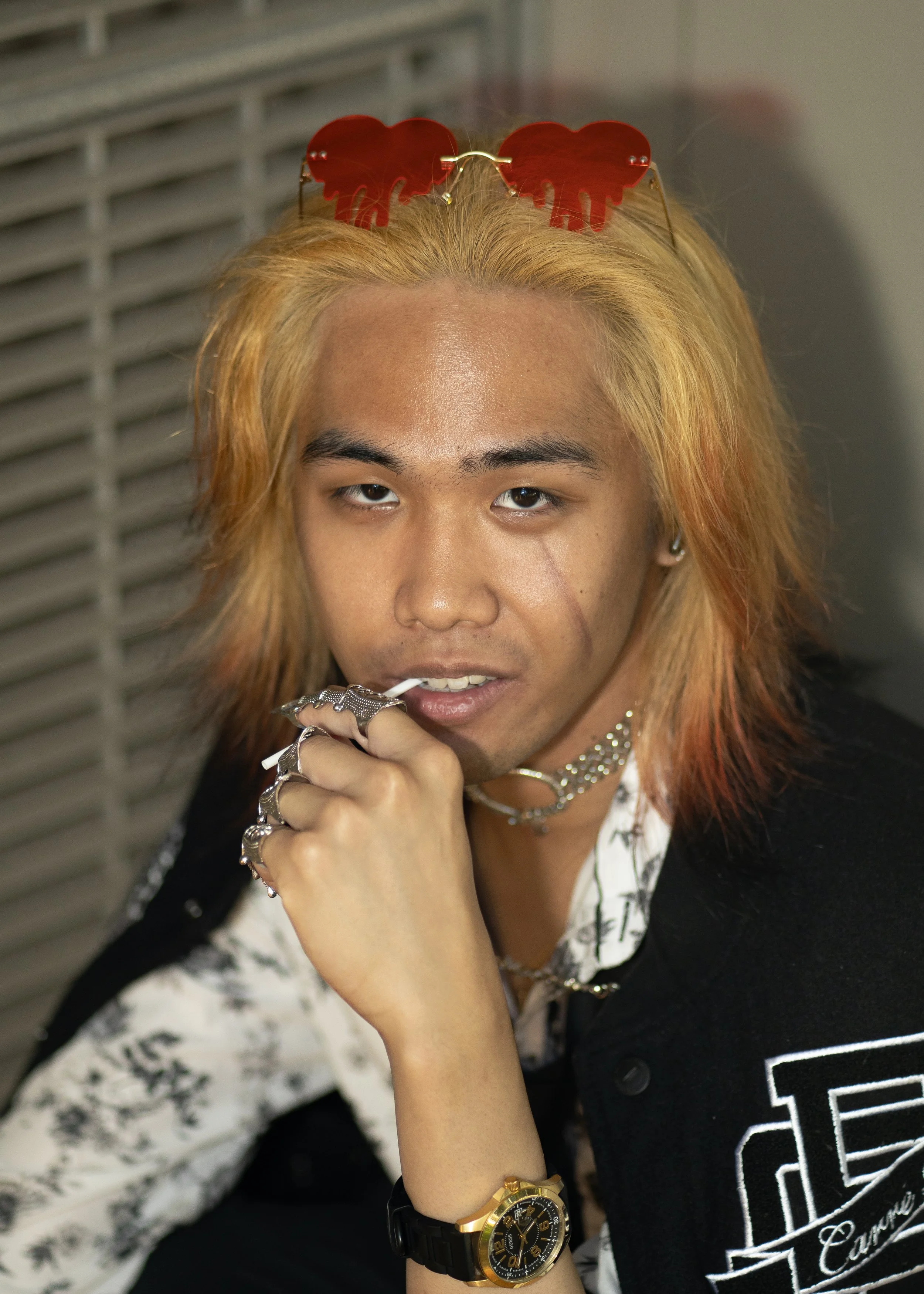 A person with blonde hair wearing red heart-shaped sunglasses, a black and white patterned shirt, layered jewelry including rings, a necklace, and a gold watch, resting their chin on their hand, with an air vent in the background.