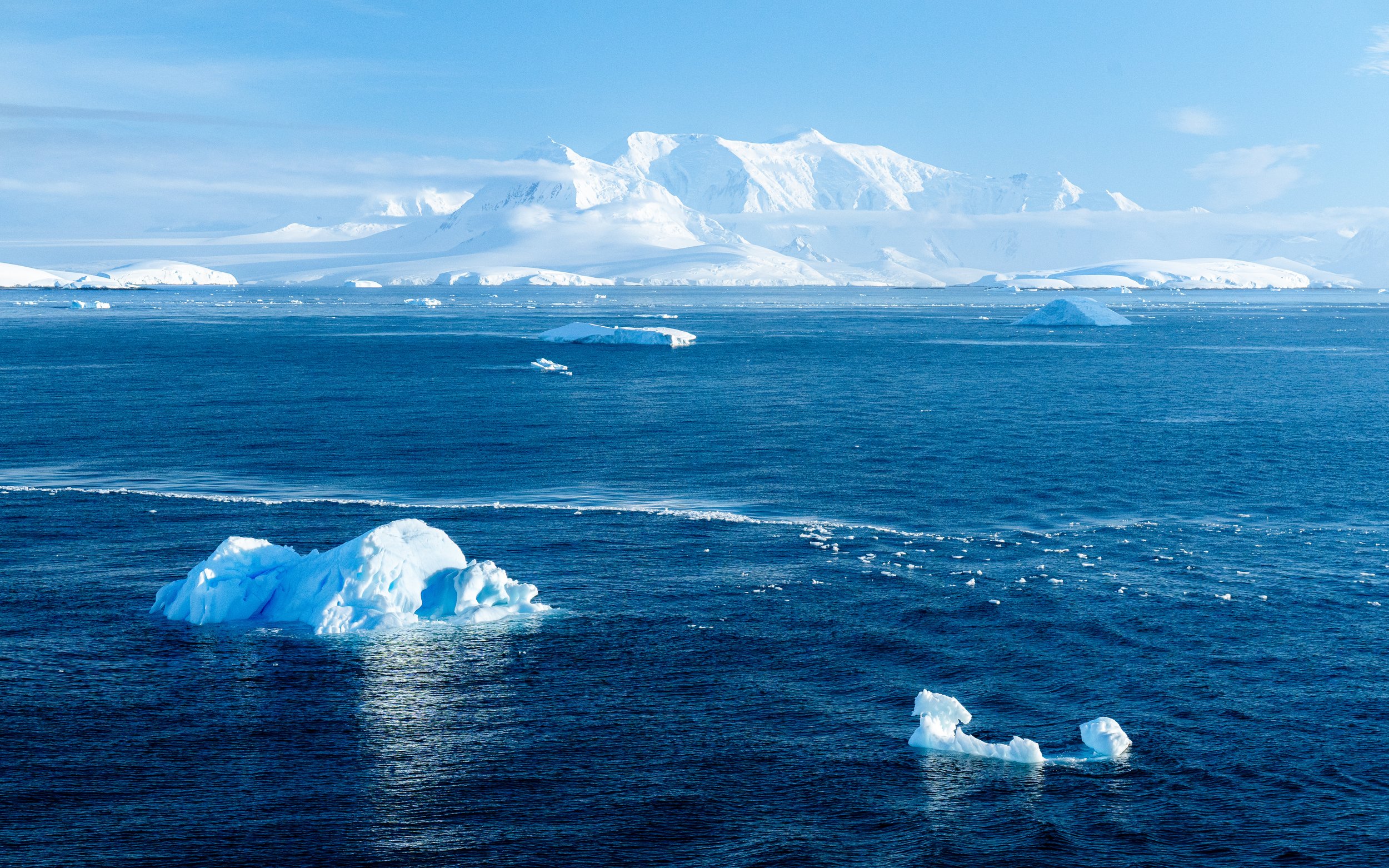 A vast icy ocean with floating ice chunks, snow-covered mountains in the background, and a blue sky with some clouds.