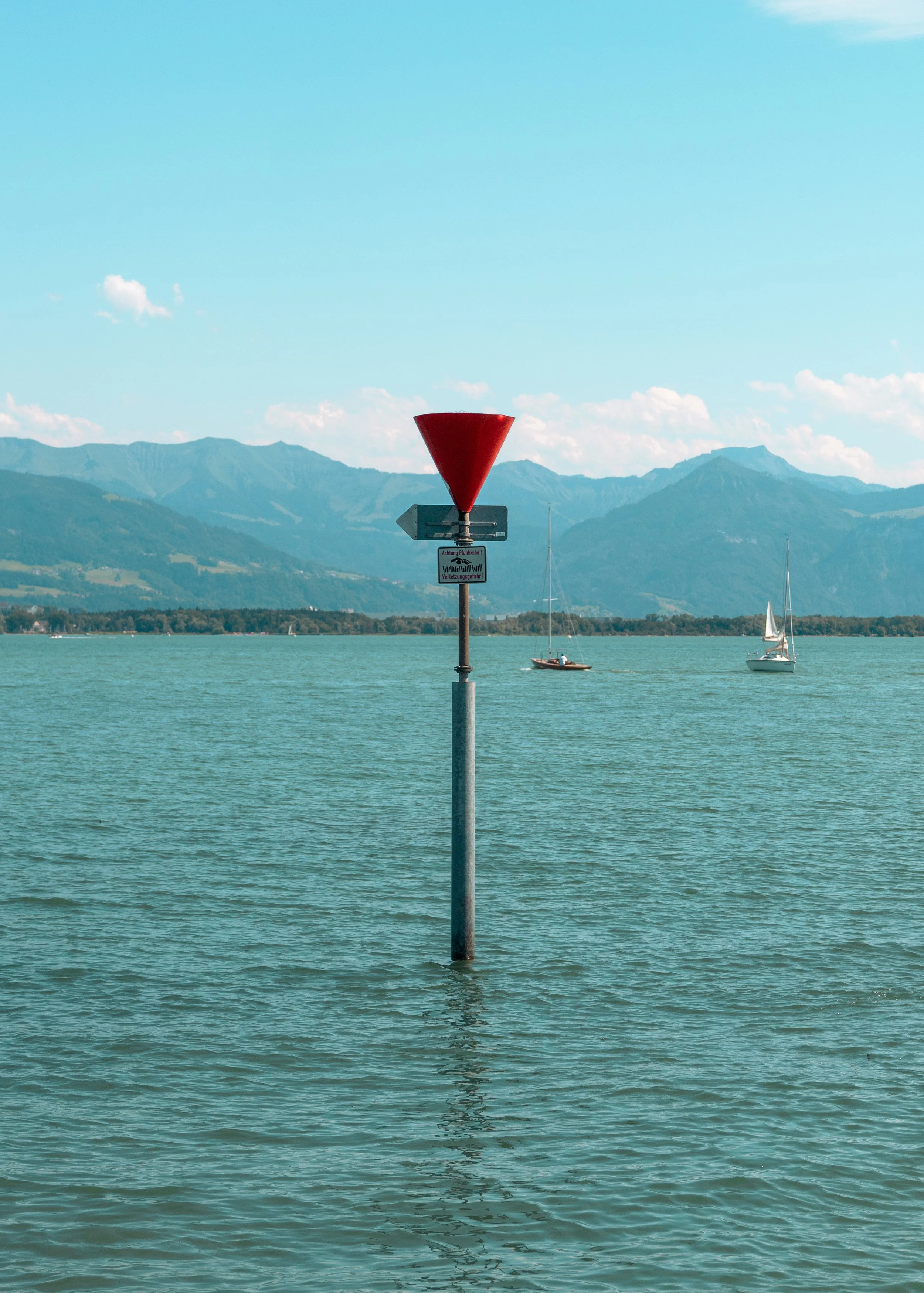 A waterway with two sailboats in the distance, a mountain range in the background, and a red navigation marker pole in the foreground.