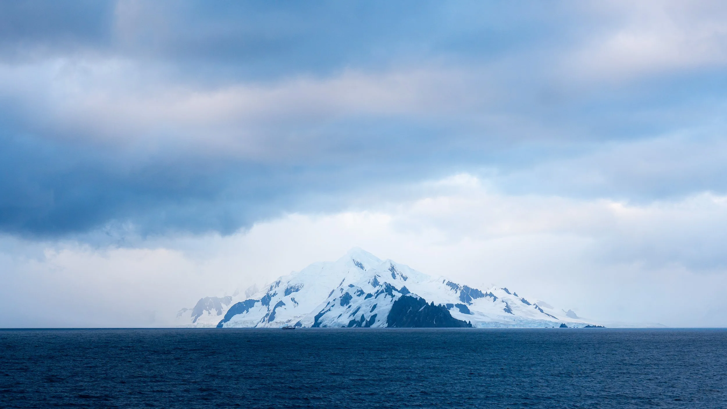 Snow-covered mountain under cloudy sky over dark ocean.