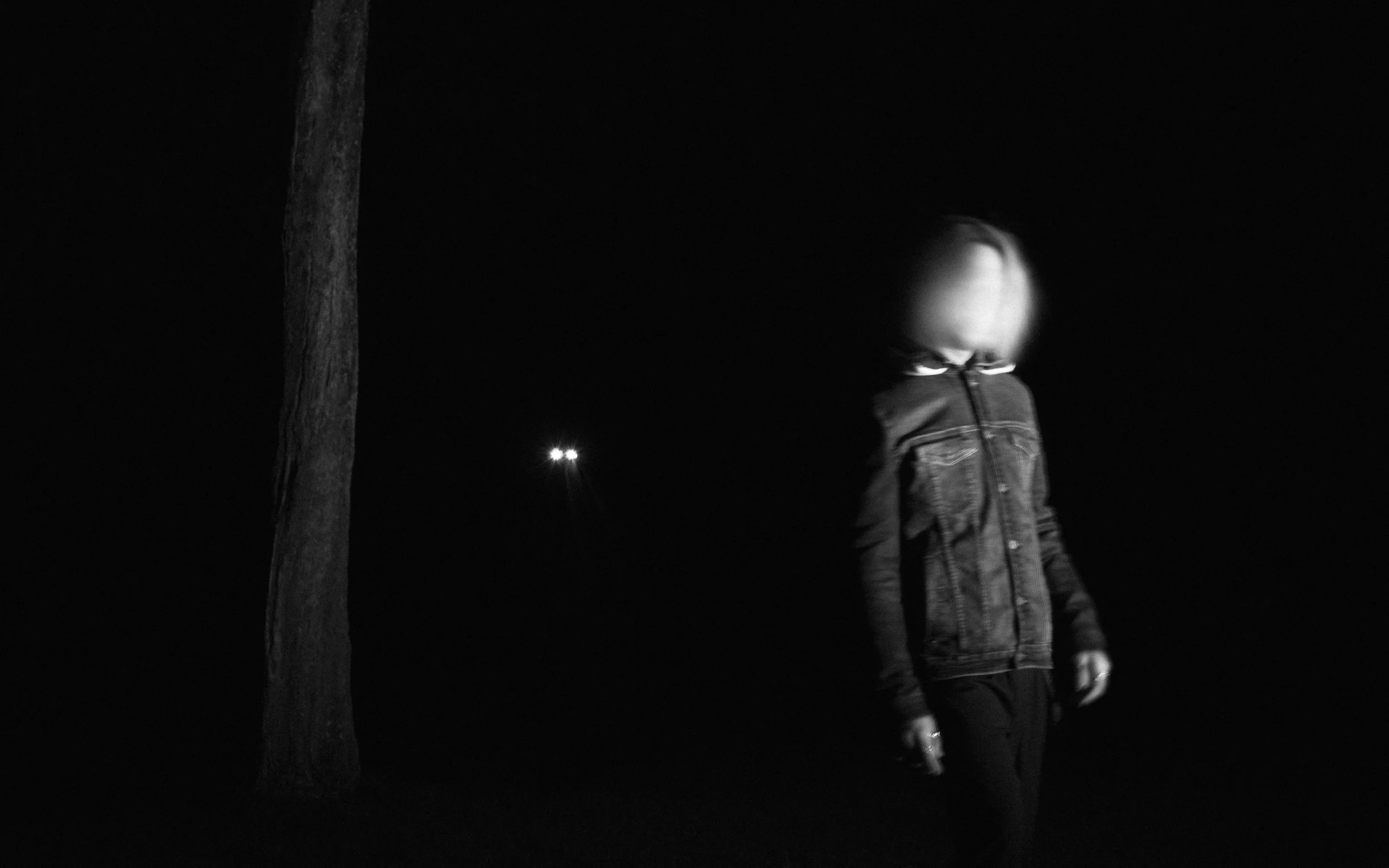 A person in a denim jacket standing outdoors at night, looking down, with a tall tree and a faint streetlight in the background.