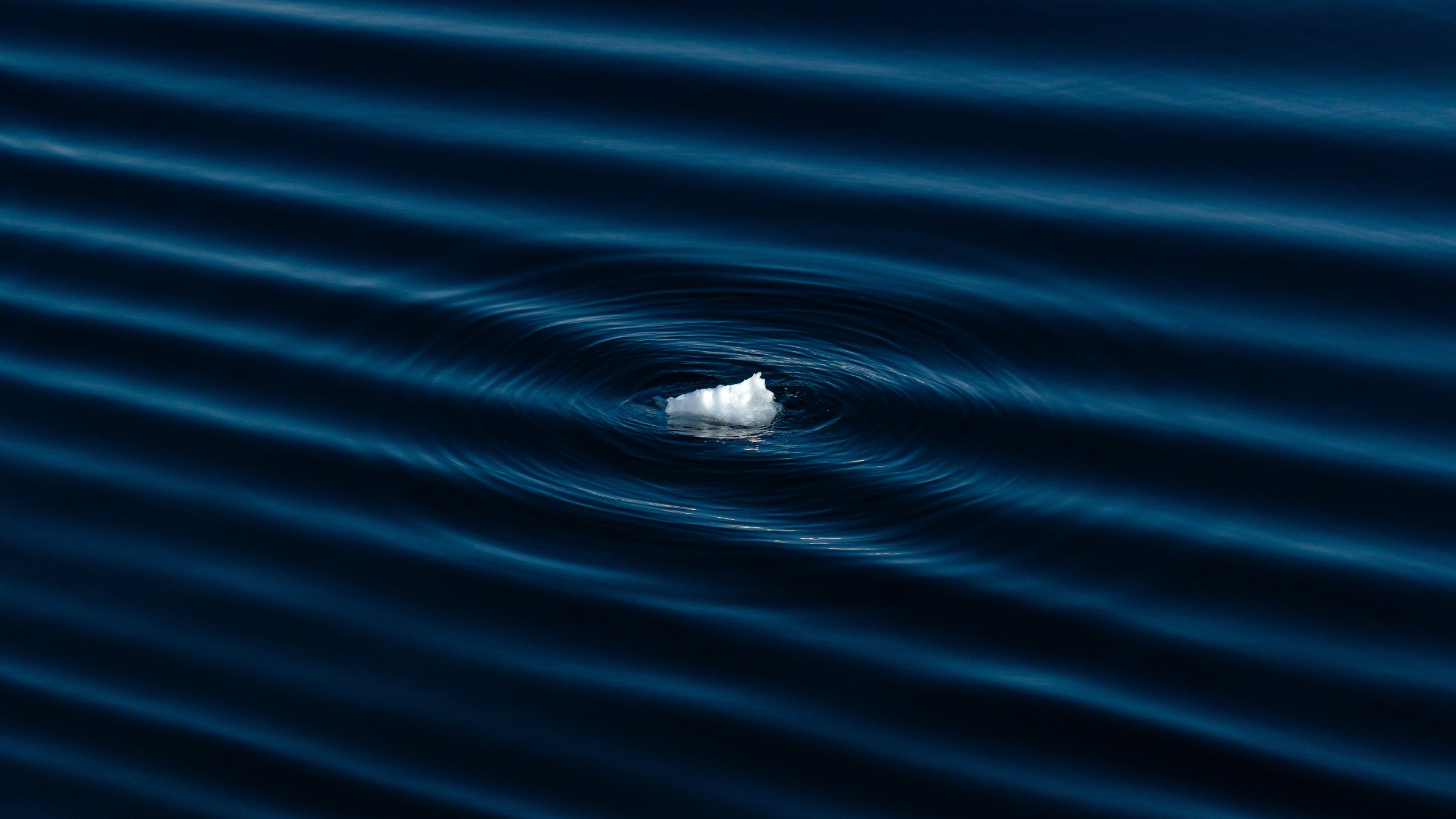 A small iceberg floating in dark blue ocean water, creating ripples as it drifts.