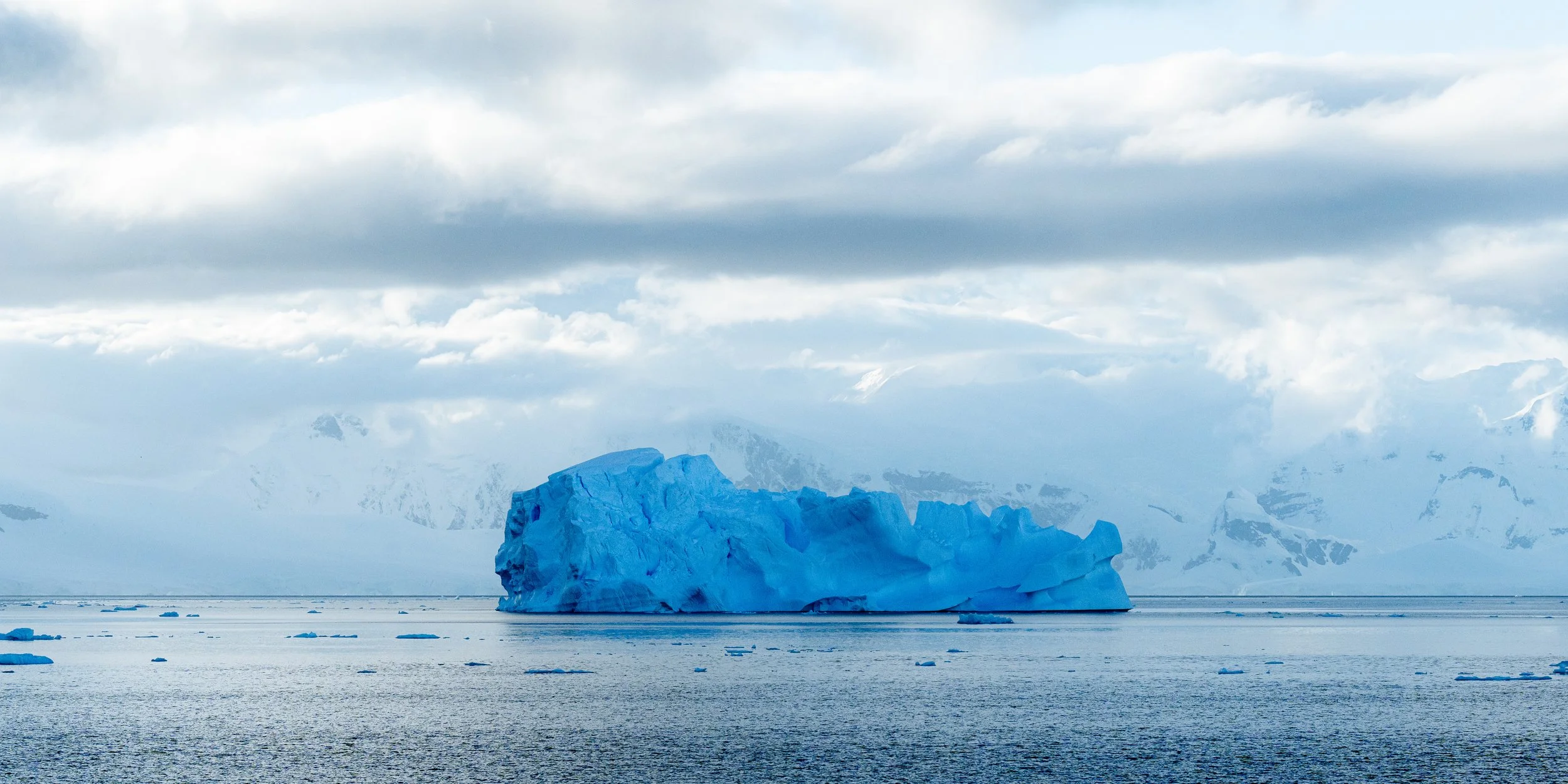 A large iceberg floating in icy waters with snow-covered mountains and clouds in the background.
