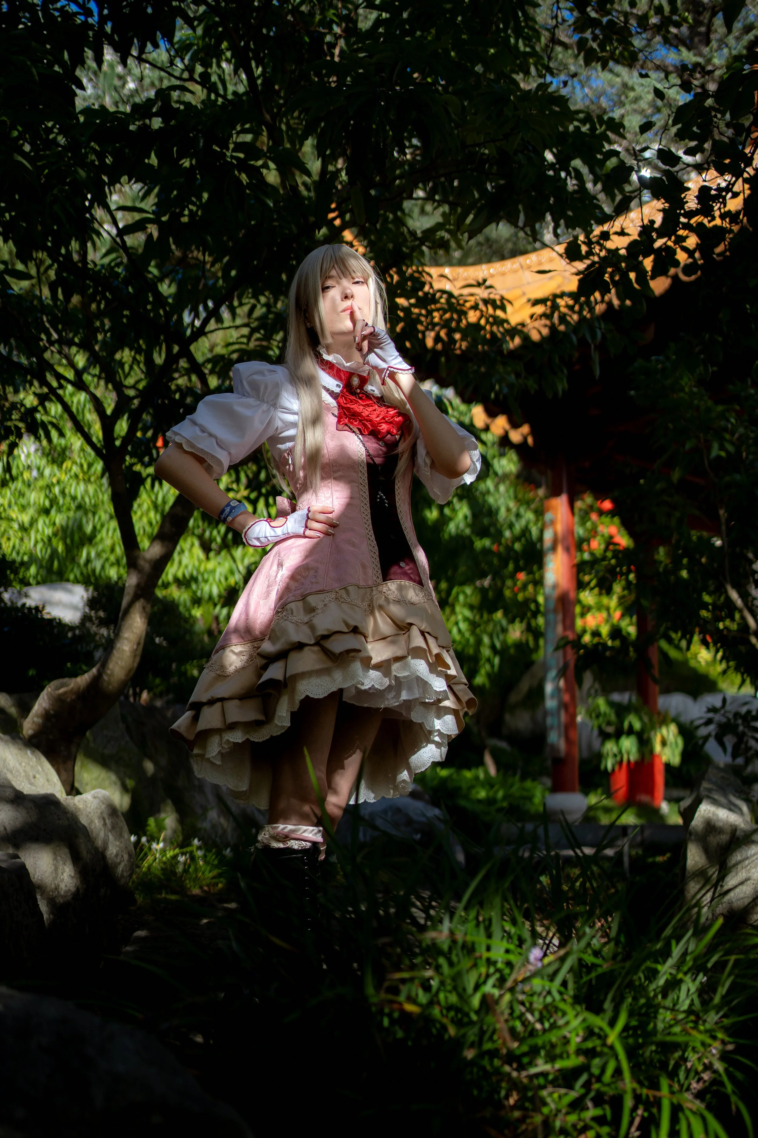 A young woman dressed in a Victorian-style pink and beige dress, standing outdoors among trees and greenery, with a finger to her lips in a shushing gesture.