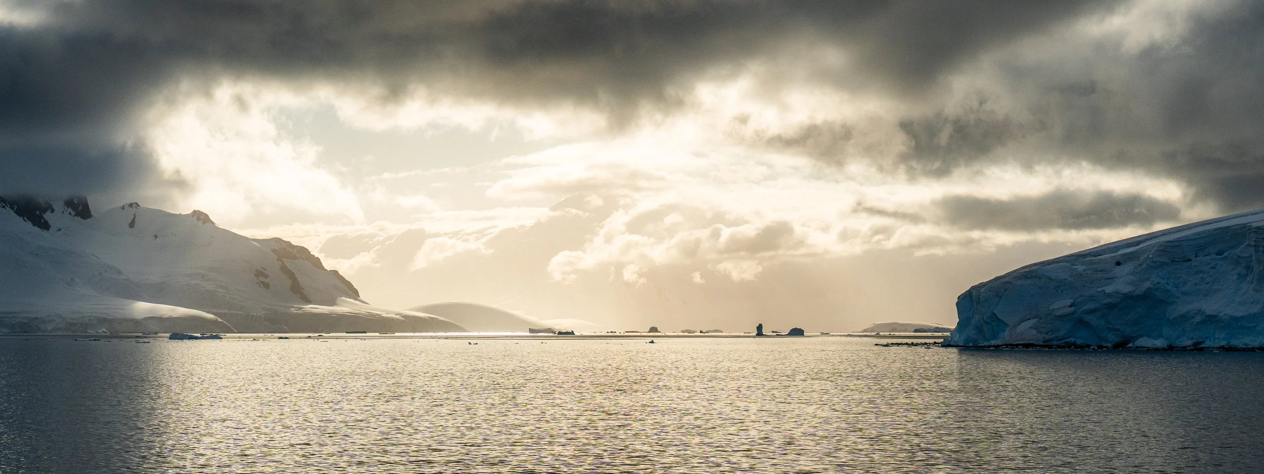 A cold, icy landscape with snow-covered mountains and large icebergs floating in the water under a cloudy sky.