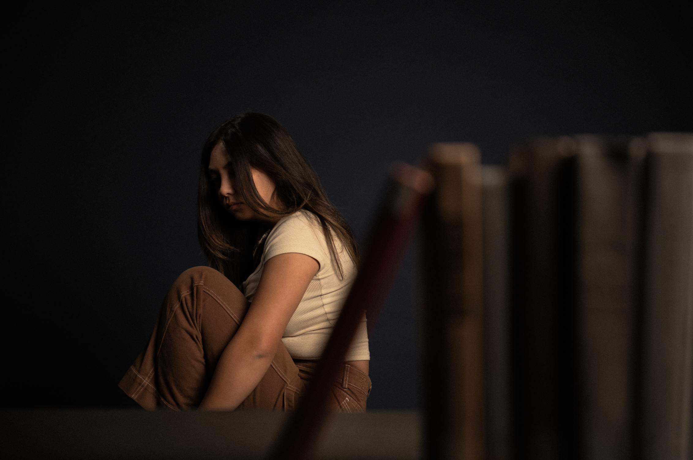 A young woman with long dark hair facing downward, seated with her arms wrapped around her knees, wearing a beige shirt and brown pants, against a dark background.
