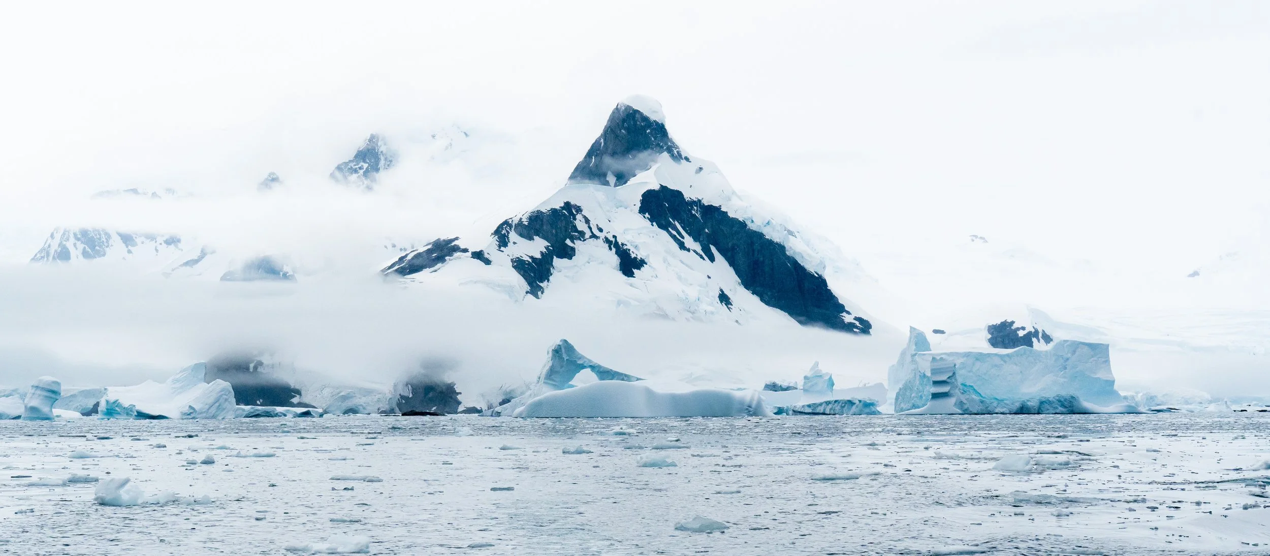 A snowy mountain peak with ice and snow-covered slopes rises above a body of water with floating ice chunks, surrounded by an overcast sky and mist.