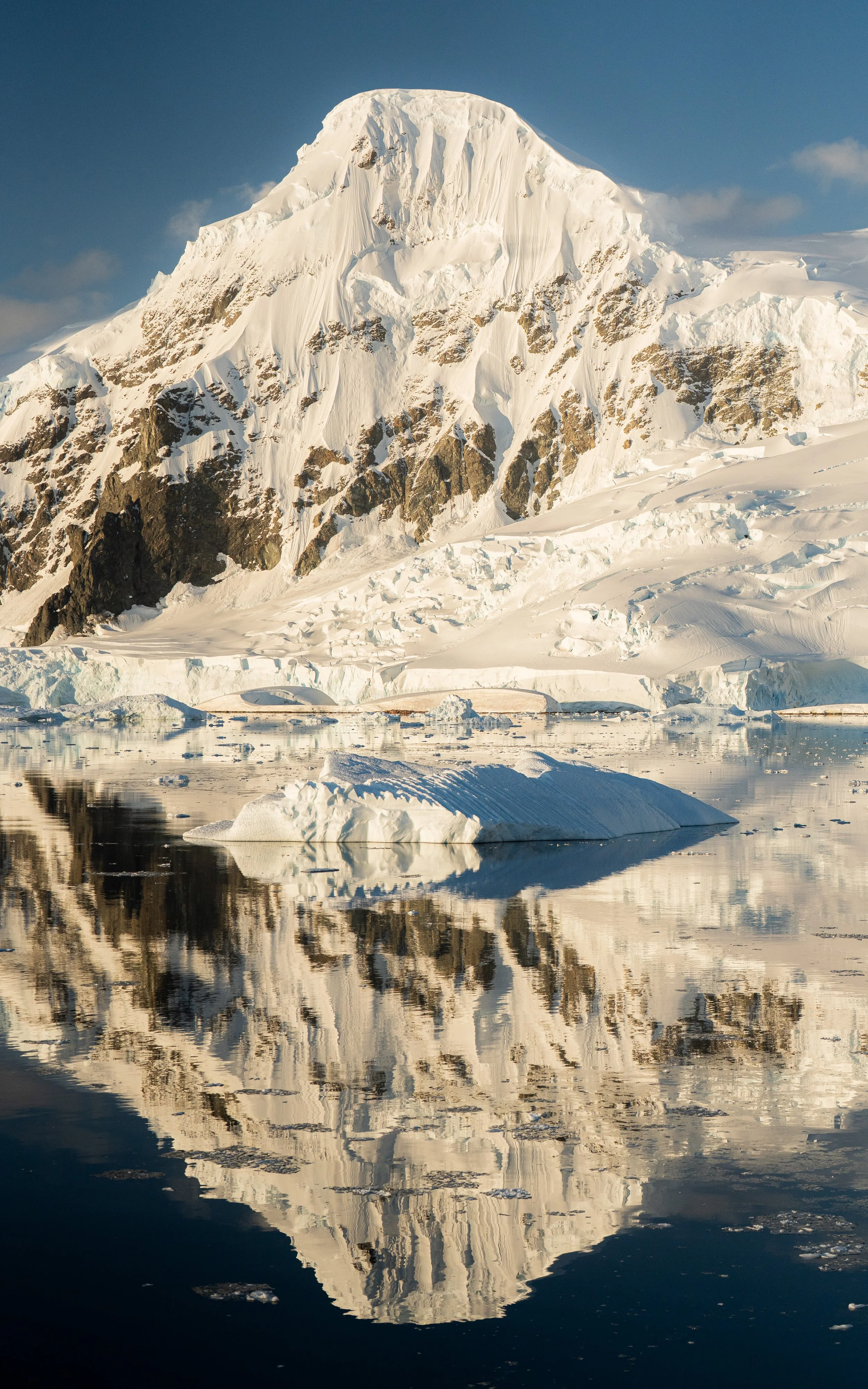 A large snow-capped mountain reflected in calm water with floating ice chunks, under a partly cloudy sky.