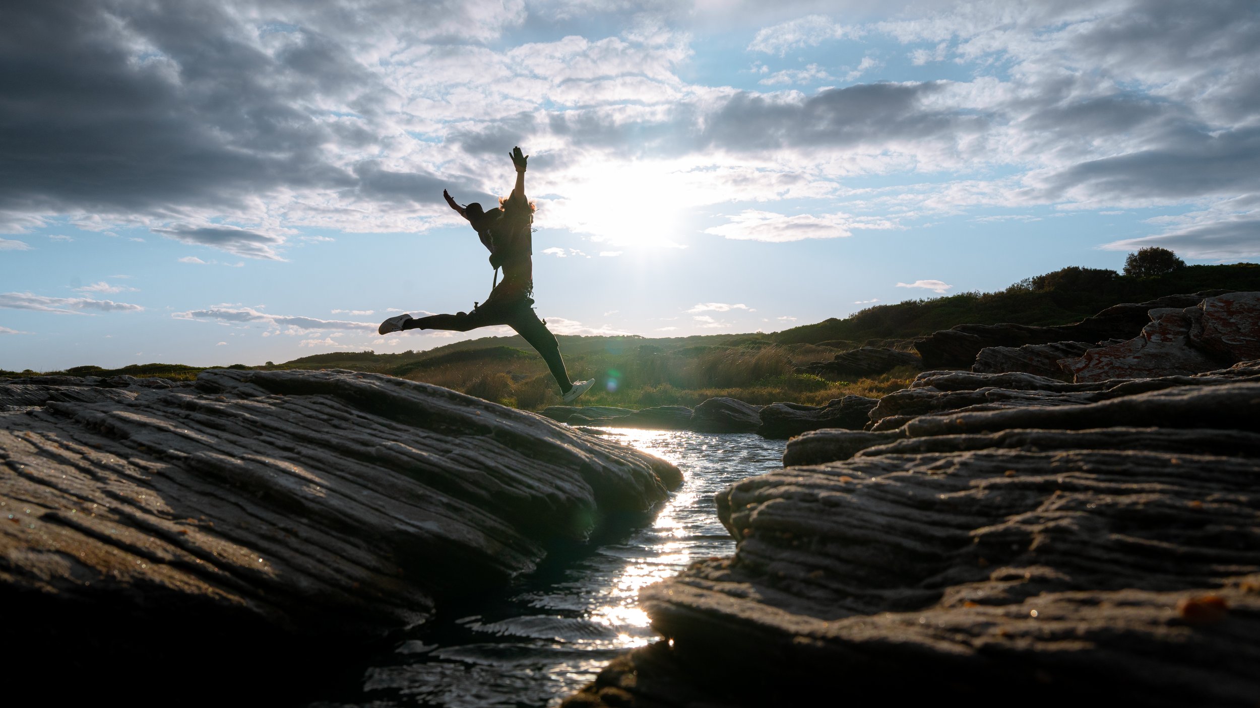 Person jumping off rocks into water during sunset with cloudy sky in the background.
