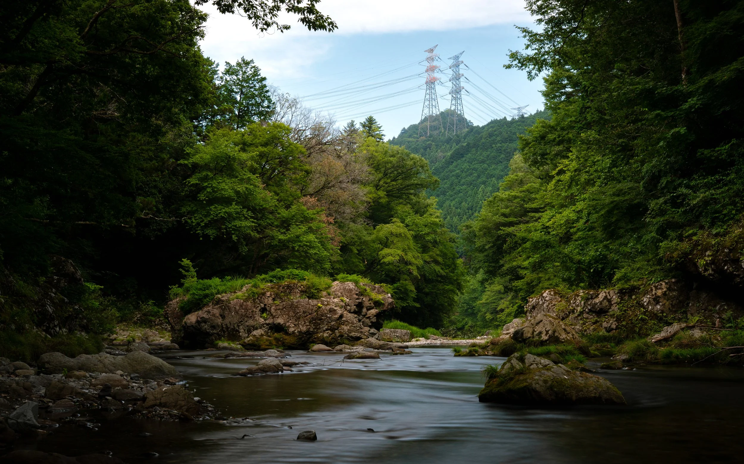 A river flowing through a lush green forested valley with rocky banks and large rocks in the water, surrounded by dense trees and distant hills, with transmission towers on the hilltop in the background.