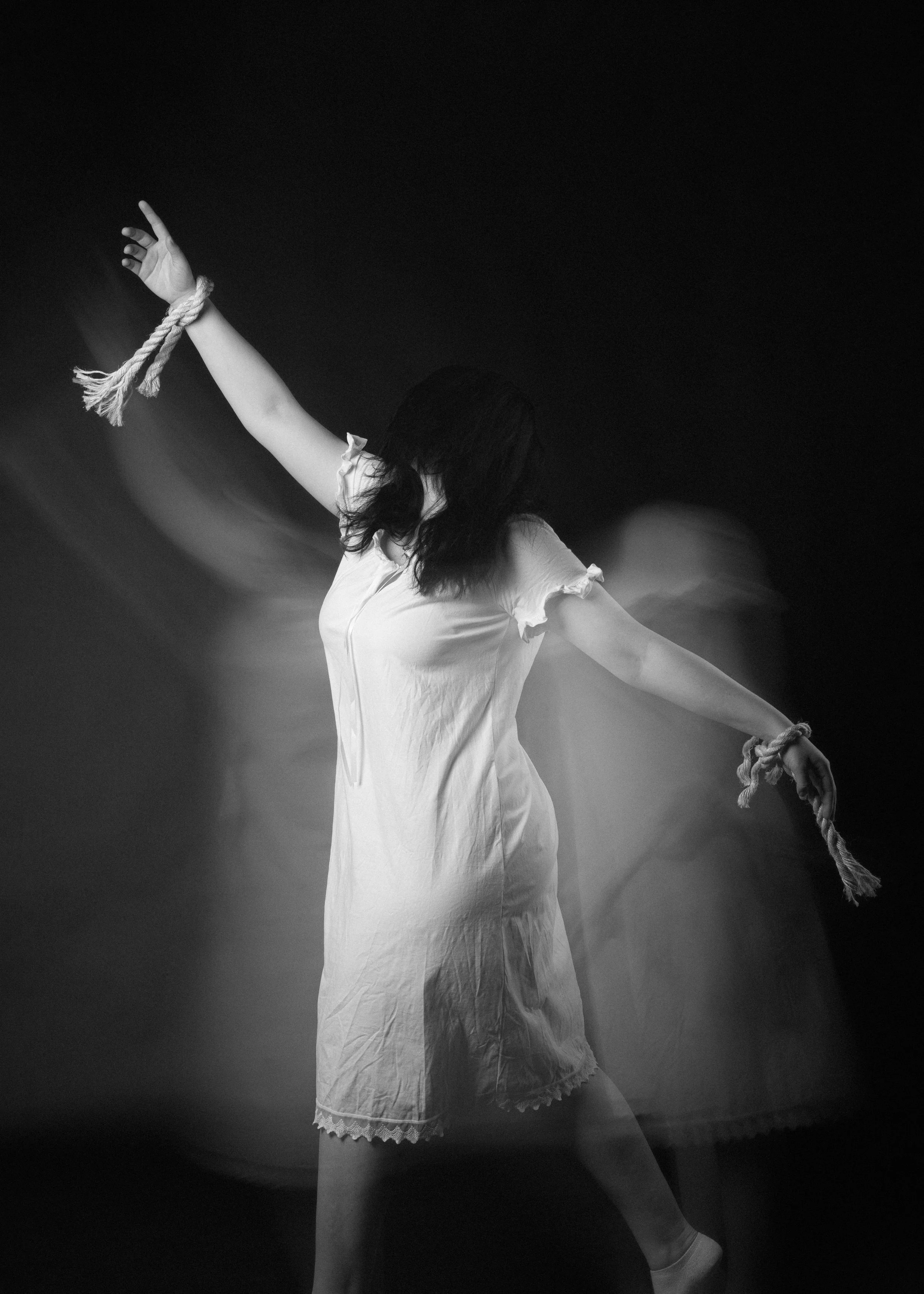 Black and white photo of a woman in a white dress with lace trim, with arms extended and tied with rope, standing on one leg with the other leg bent, in a dark, indistinct background.