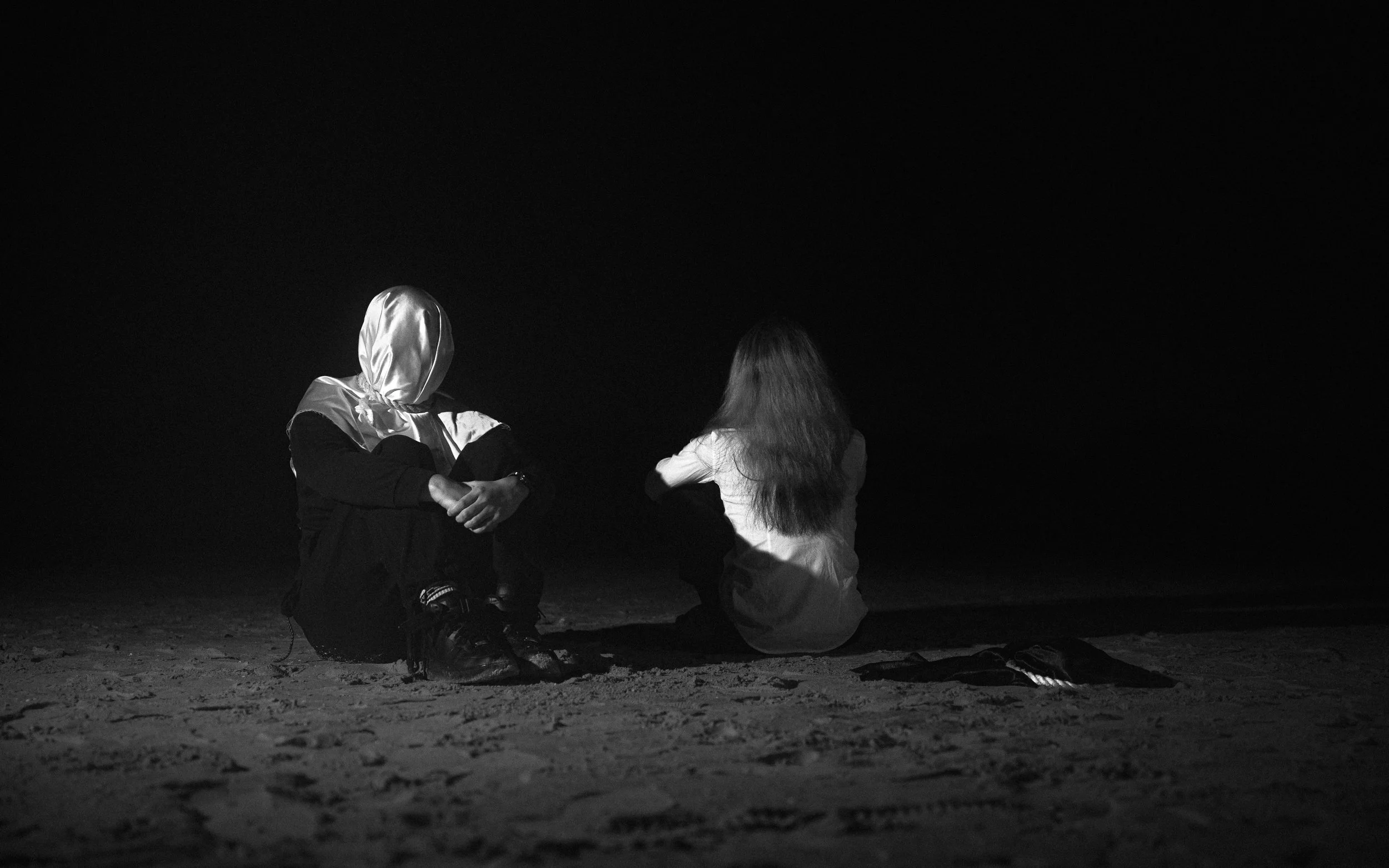 Two people are sitting on the ground at night, one wearing a shiny hood and the other with long hair, against a dark background.