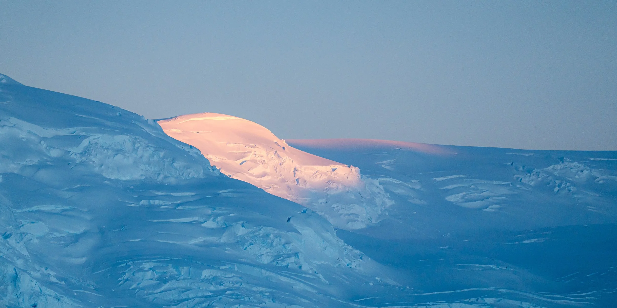 Snow-covered landscape with icy mountains under a clear blue sky, illuminated by soft pink sunlight.