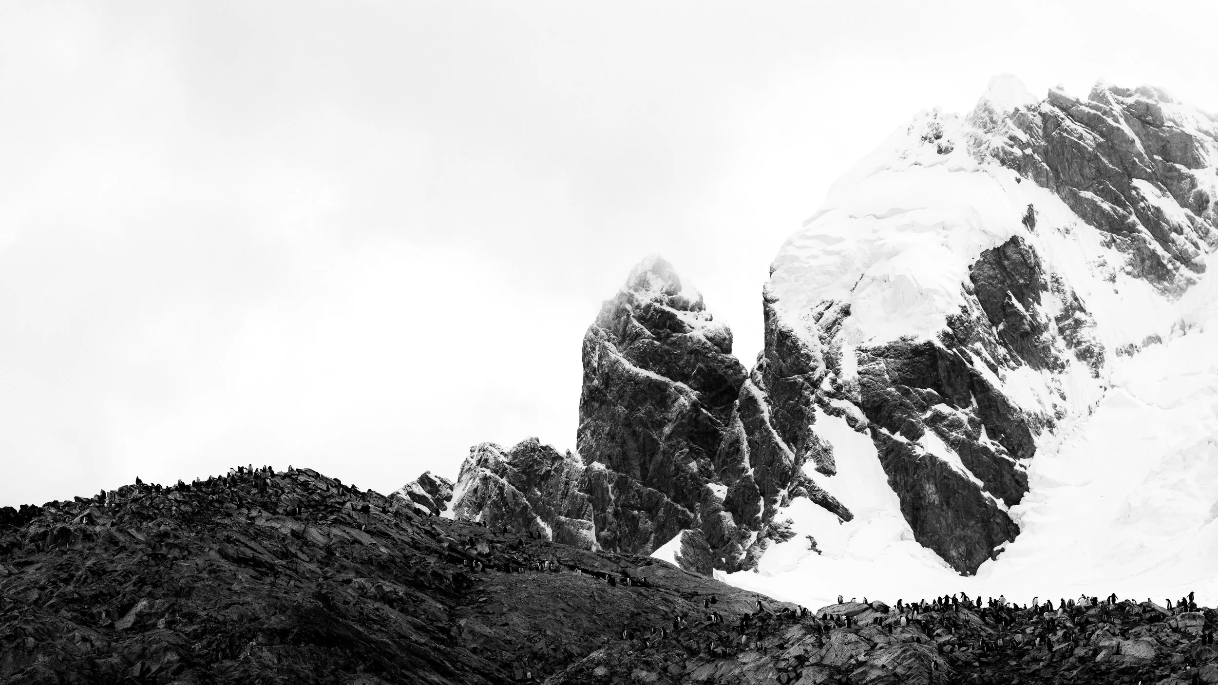 Black and white photograph of a snow-covered mountain with a steep rocky slope at its base, and a line of small climbers or hikers on the slope.