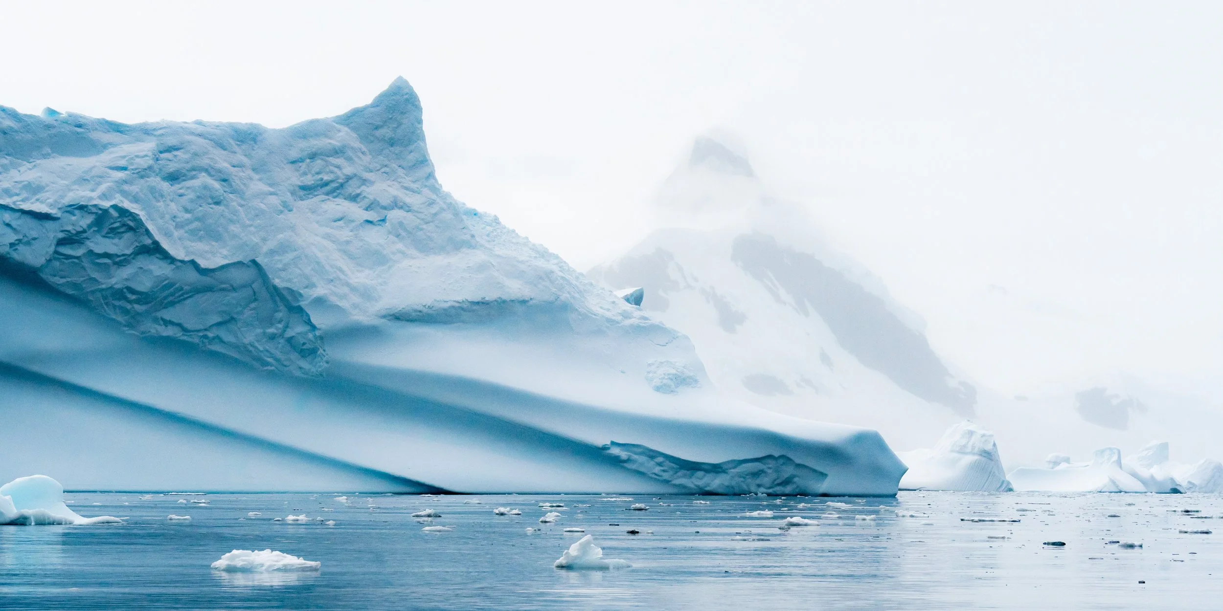 Iceberg floating in the Arctic Ocean with icy mountains in the background, under a cloudy sky.