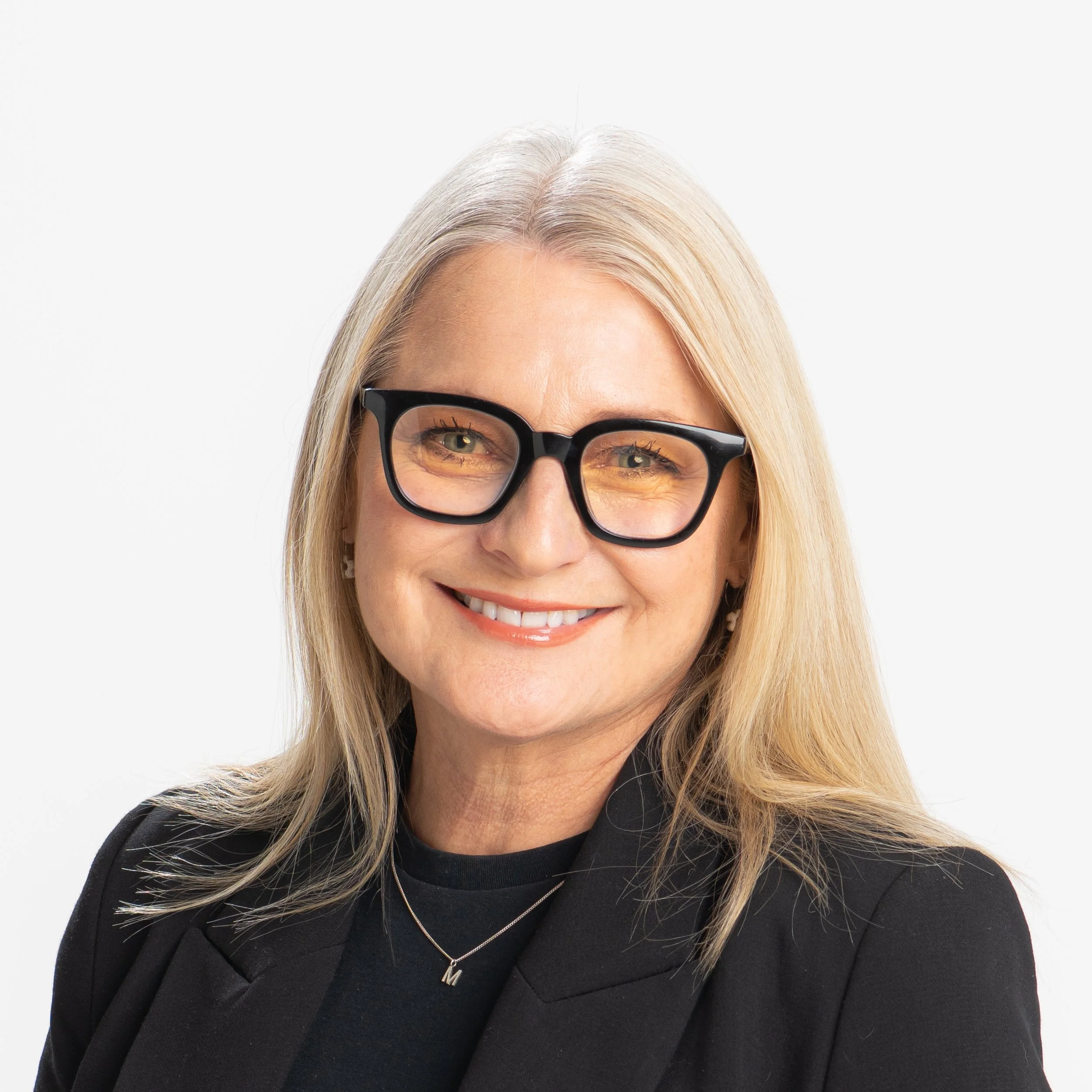 A close-up of a woman with blonde hair wearing black thick-rimmed glasses, a black blazer, and a black top, smiling against a plain white background.