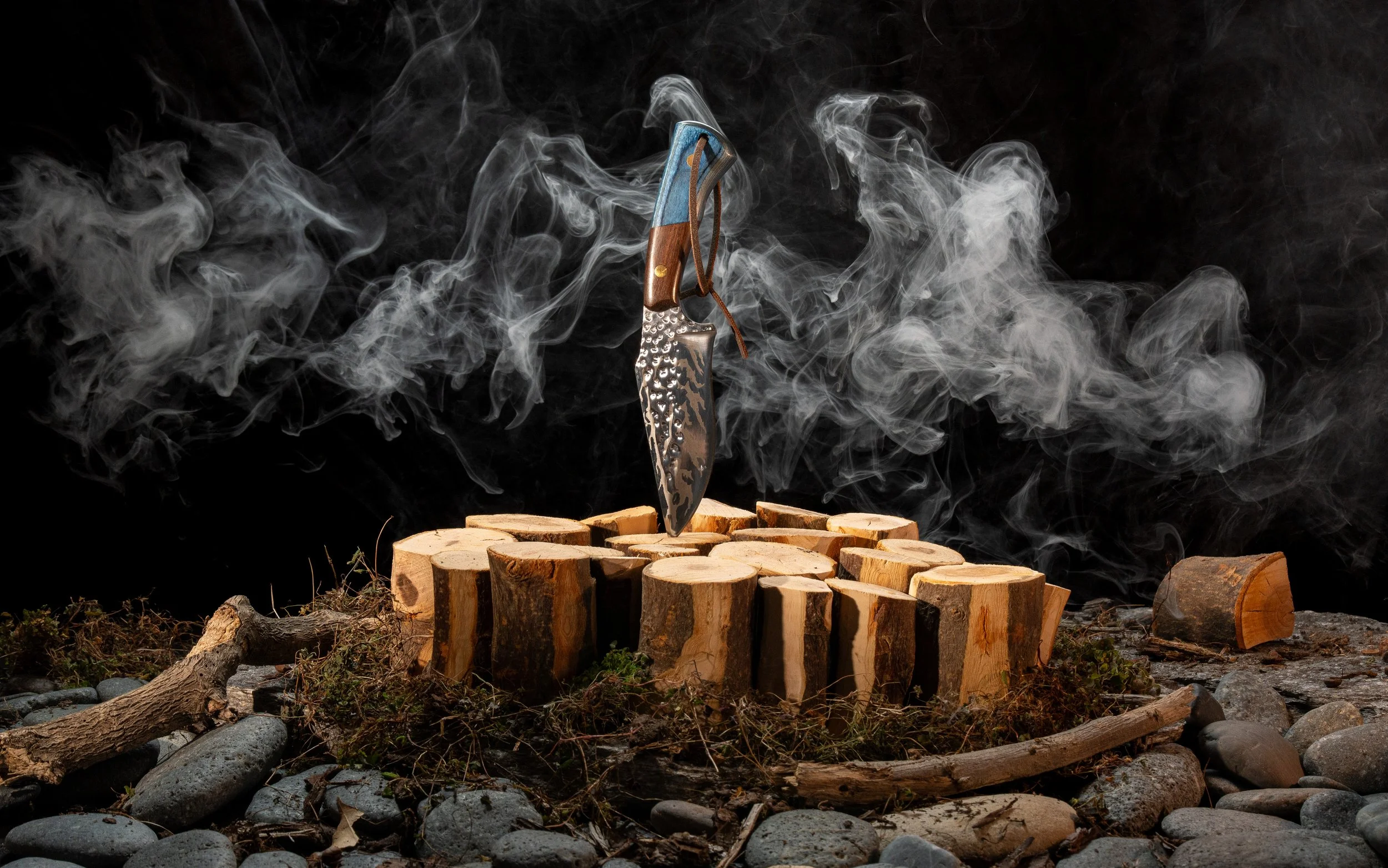 A fixed-blade knife with a textured metal blade and a handle made of wood and blue material, mounted on a wooden chopping block surrounded by stones and twigs, with smoke billowing in the background.