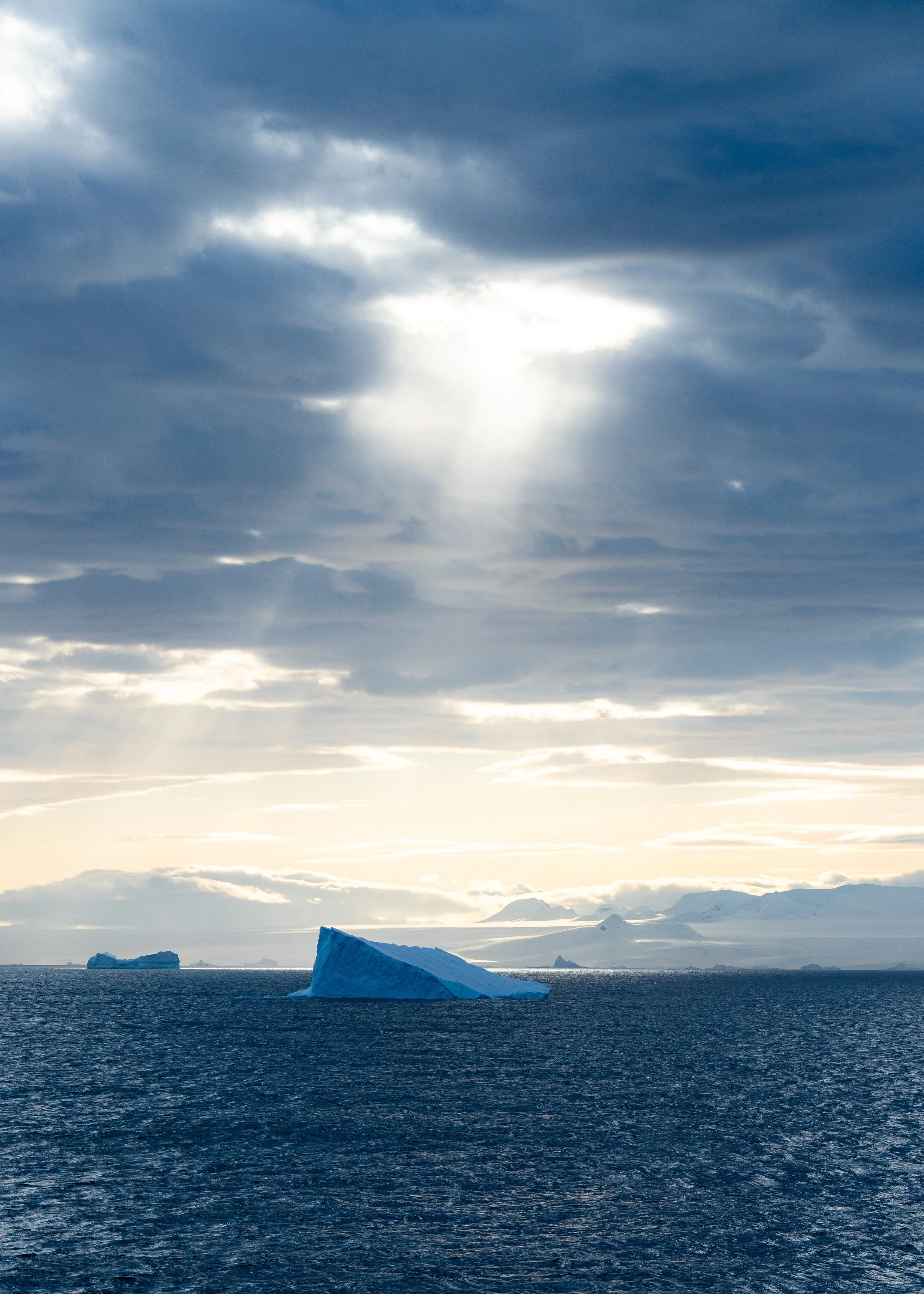A large iceberg floating in the dark ocean under a cloudy sky with sunlight shining through the clouds.