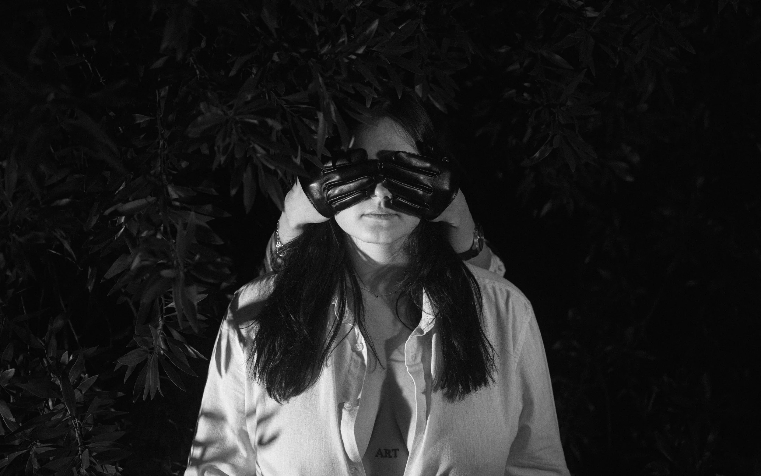 A person with long dark hair wearing a white shirt and black gloves covering their eyes with their hands, surrounded by dense dark leaves.