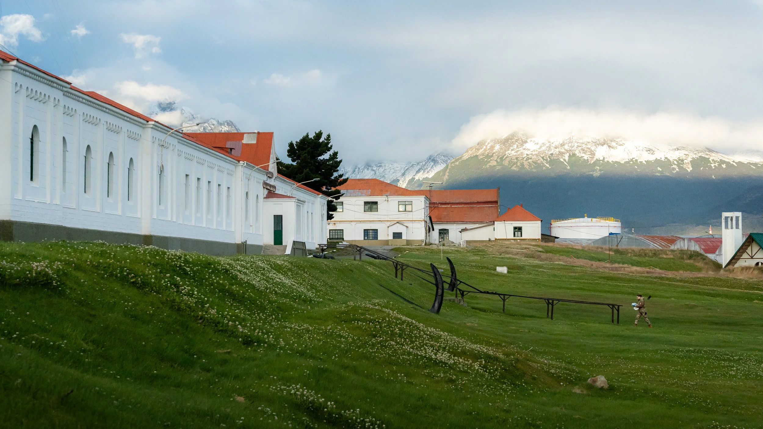 A scenic view of a small town with white buildings and red roofs, set against tall snow-capped mountains partly covered by clouds, with a grassy foreground and a person sitting on a bench.