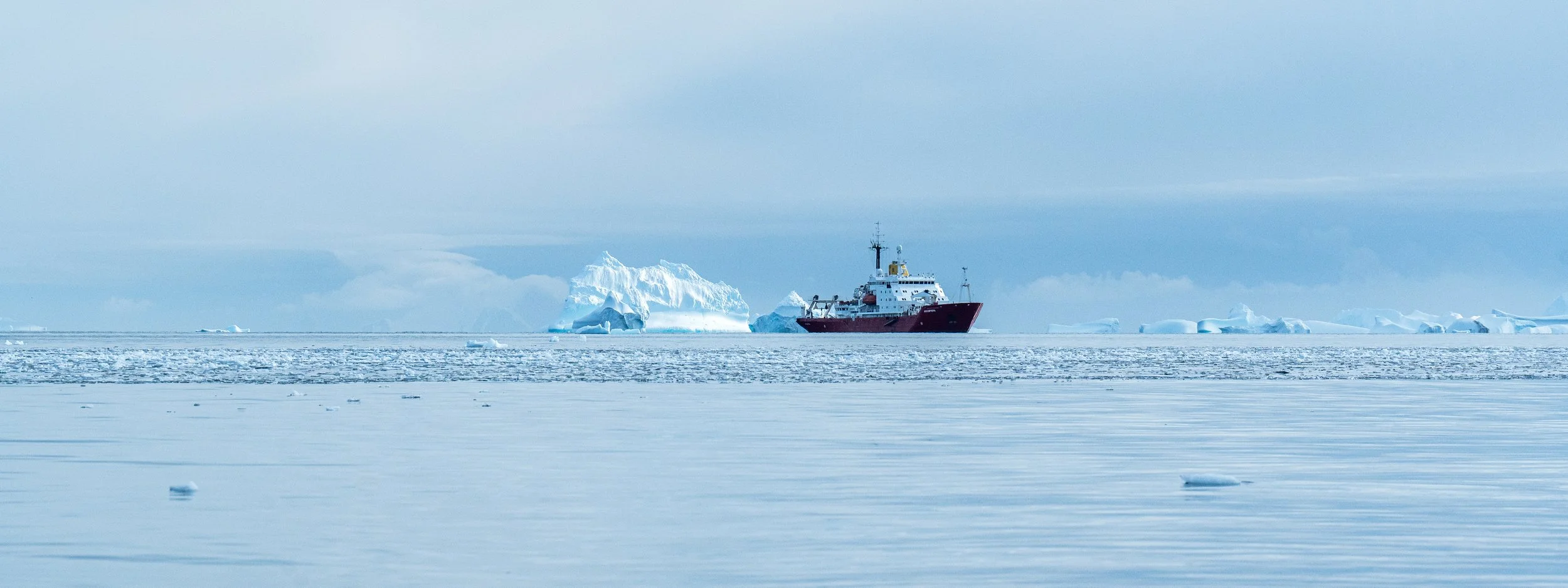 A red and white ship sailing through icy waters with large icebergs in the background under a cloudy sky.