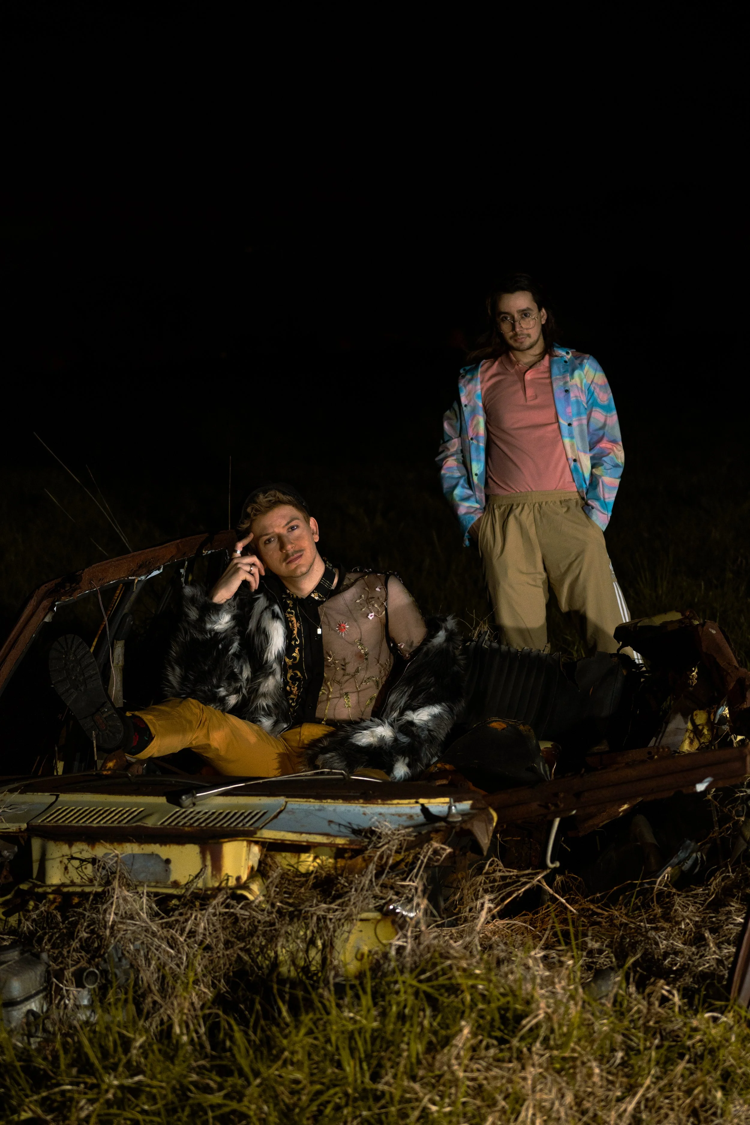 Two young men with long hair, one wearing glasses, posing near a rusted, abandoned car in a field at night. One sitting inside the car, the other standing beside it, both dressed in casual clothing.