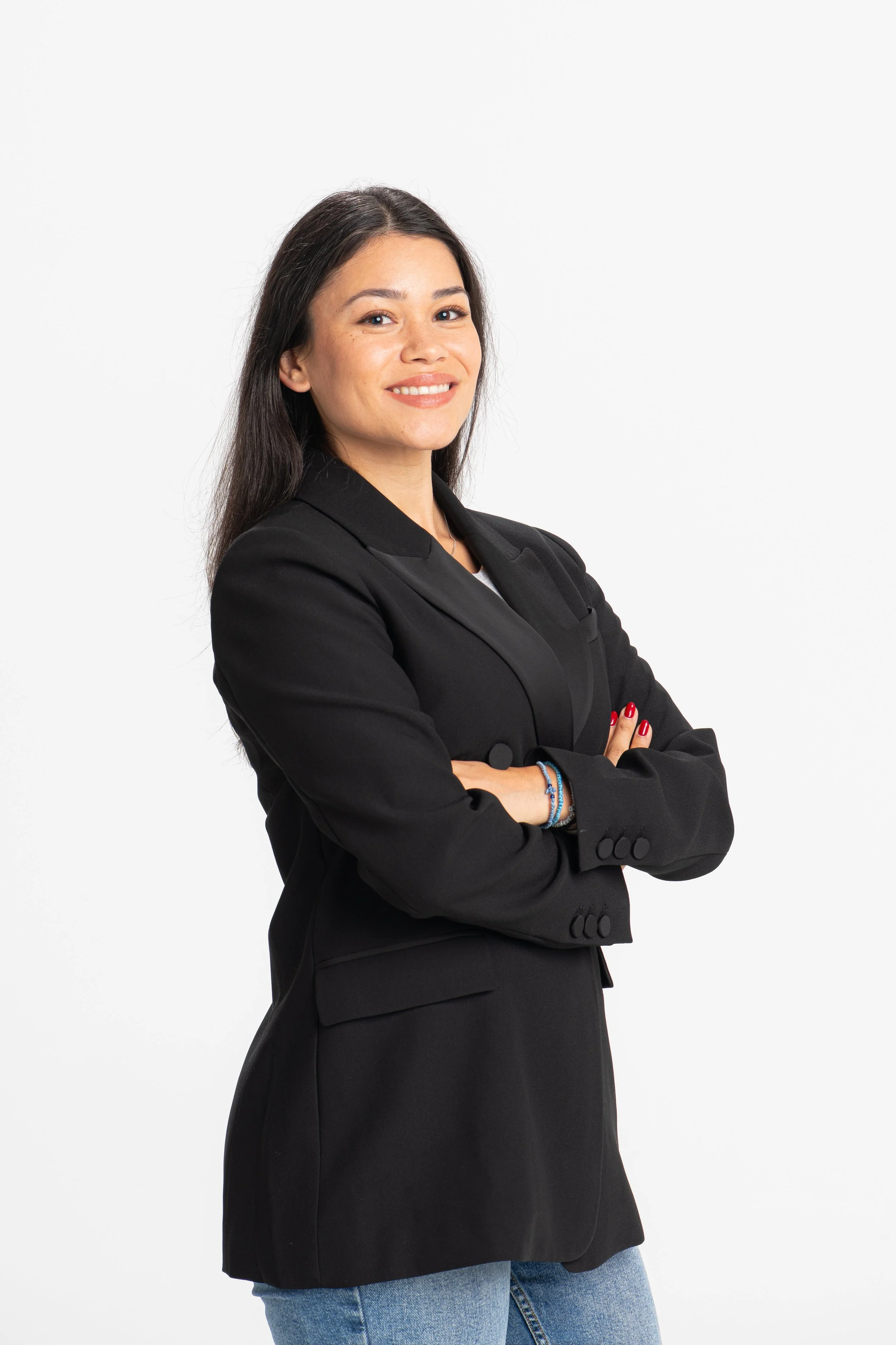 A woman with dark brown hair and light skin, smiling, dressed in a black blazer, white top, and jeans, standing against a plain white background.