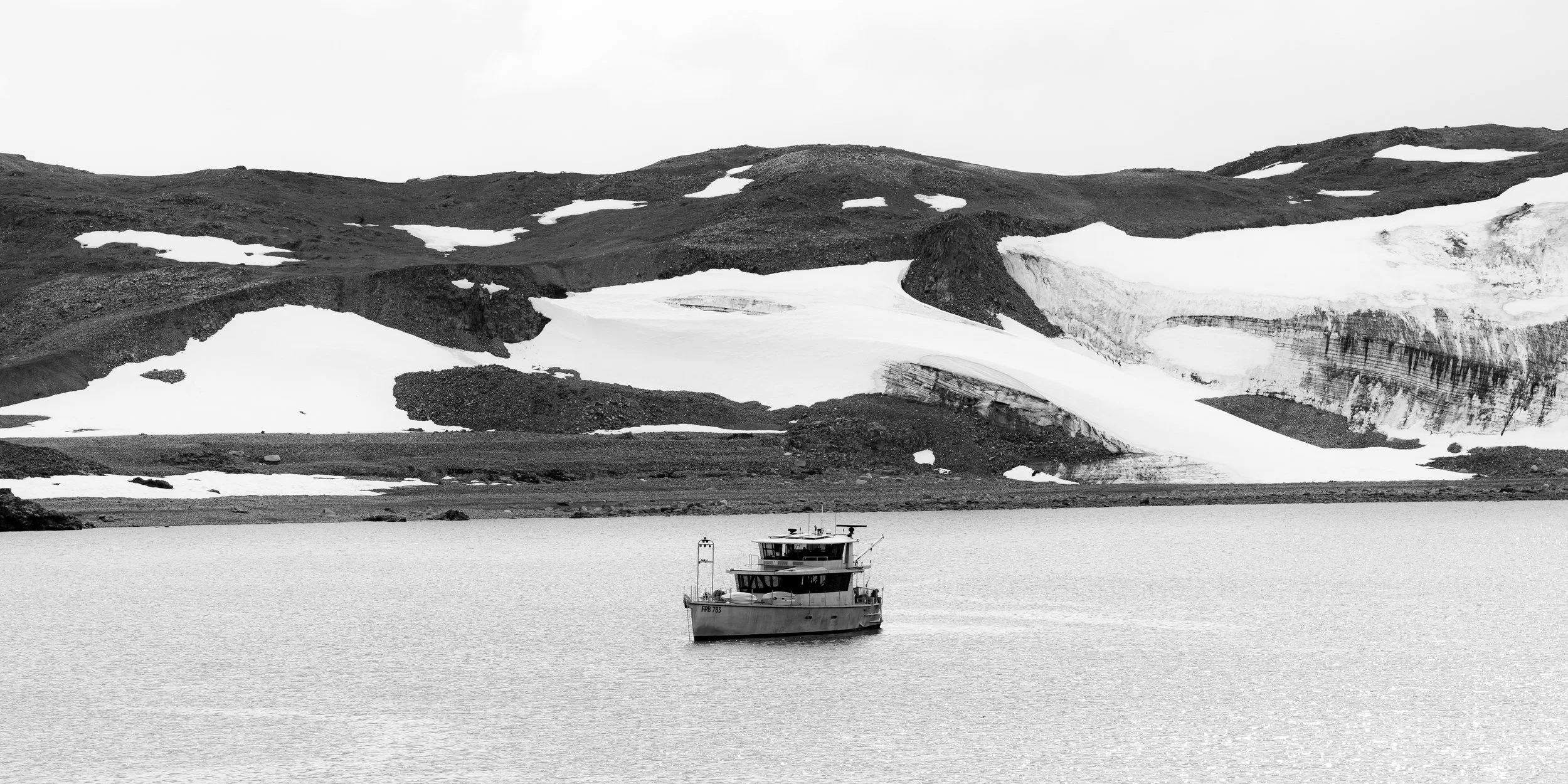 A black and white photograph of a boat on a calm body of water, with snow-covered mountains and rocky terrain in the background under an overcast sky.