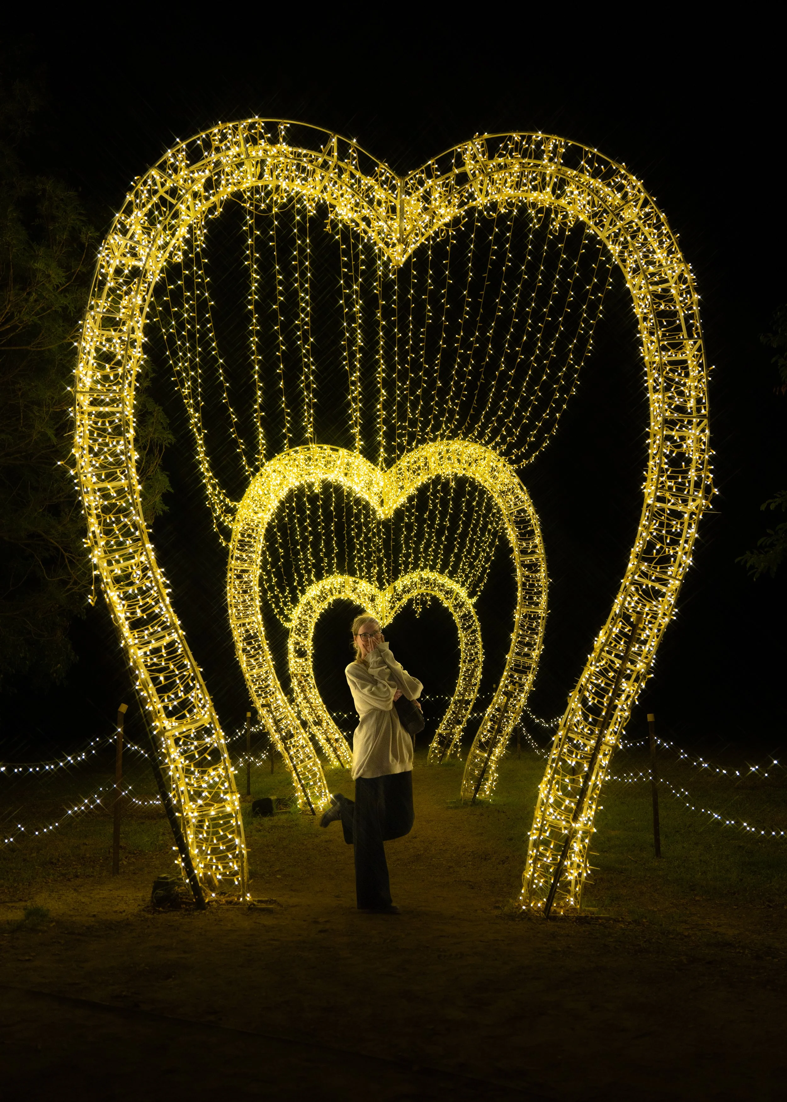 Person dressed in light-colored clothing, holding a bag, standing under illuminated heart-shaped light decorations at night.