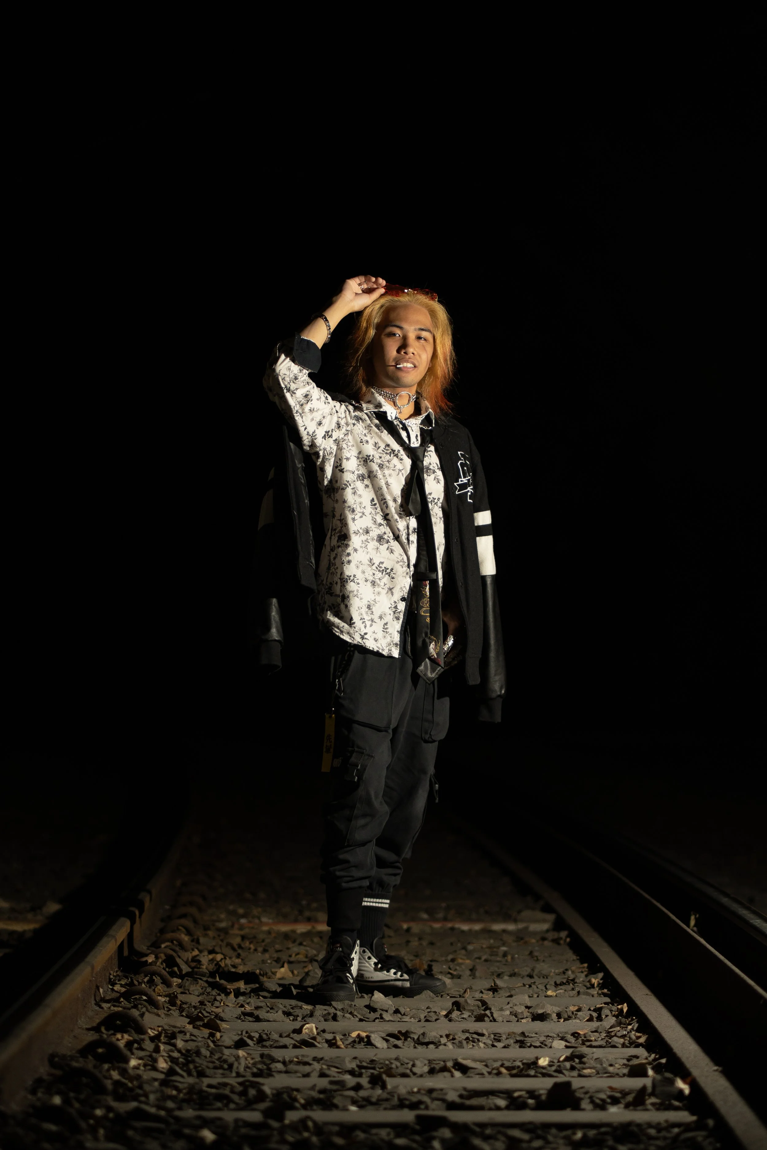 Young man with blonde hair standing on railroad tracks at night, adjusting sunglasses, wearing a patterned shirt, black jacket, cargo pants, and sneakers, with dark background.