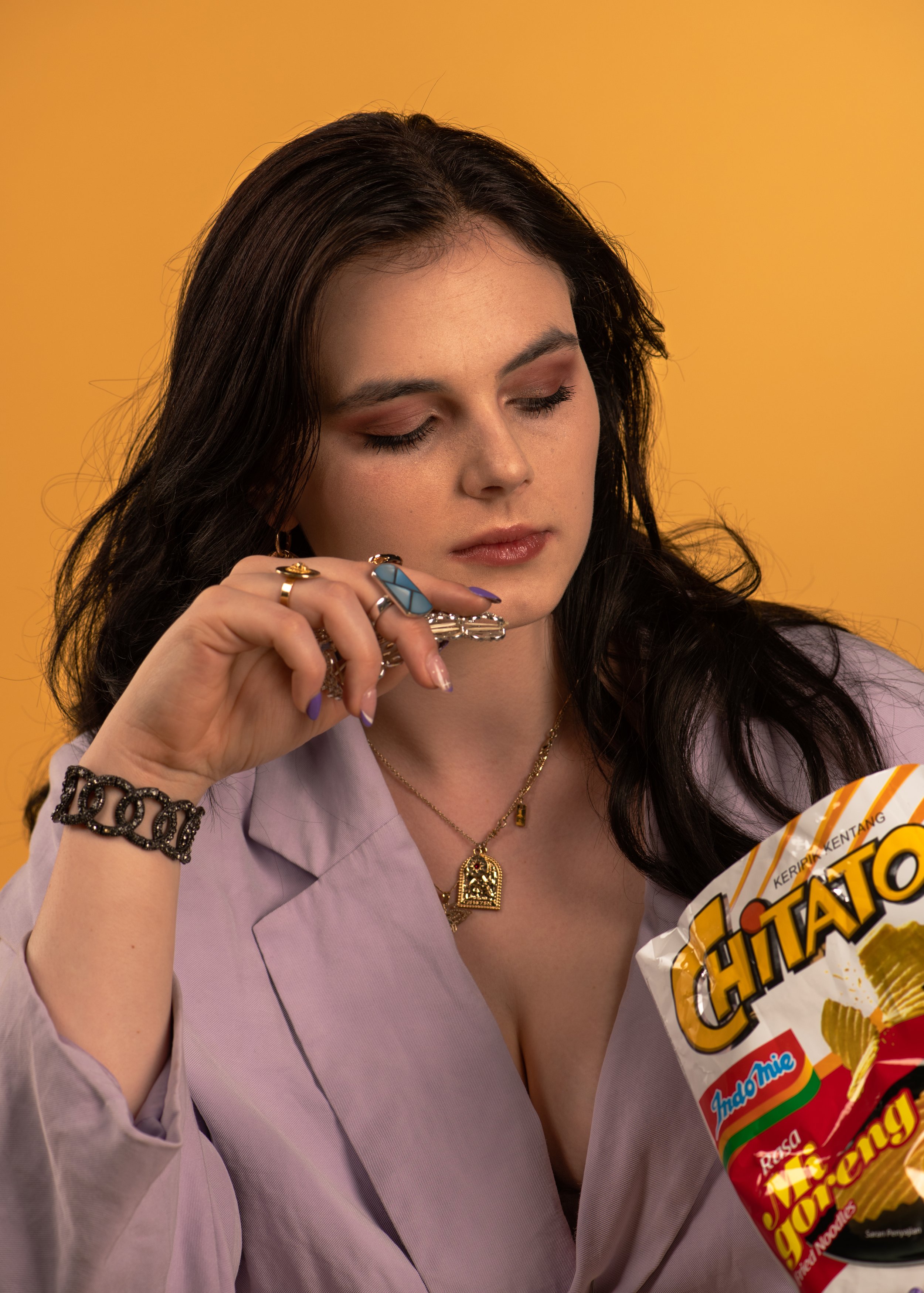 A woman with dark hair, wearing jewelry, holding a bag of Chitato potato chips, against a plain yellow background.