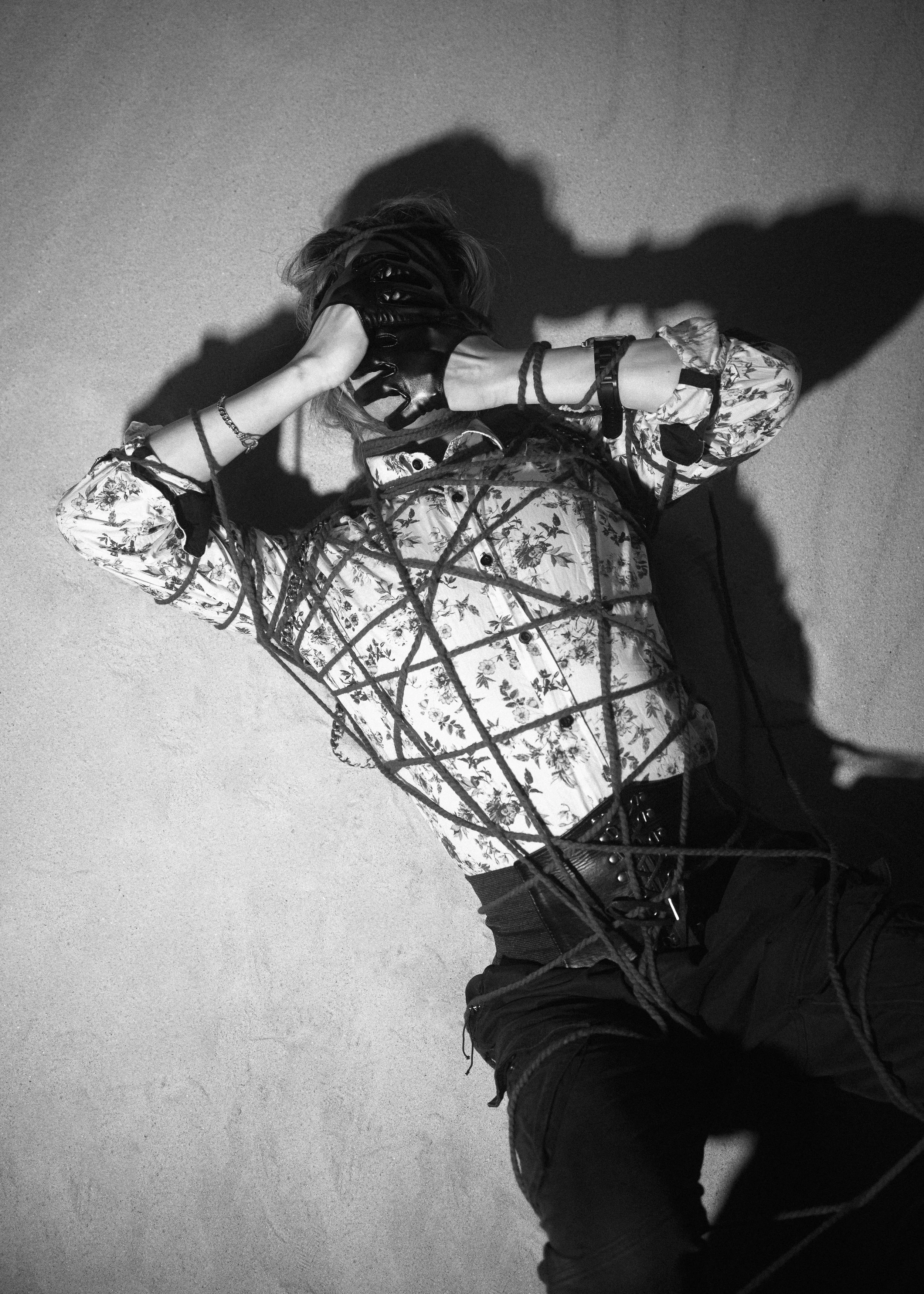 A person wearing a floral-patterned jacket and dark pants is lying on the ground. They are holding a pair of headphones or earmuffs over their ears with their right hand. Their shadow is cast on a plain, textured wall or floor, and their face is not visible.