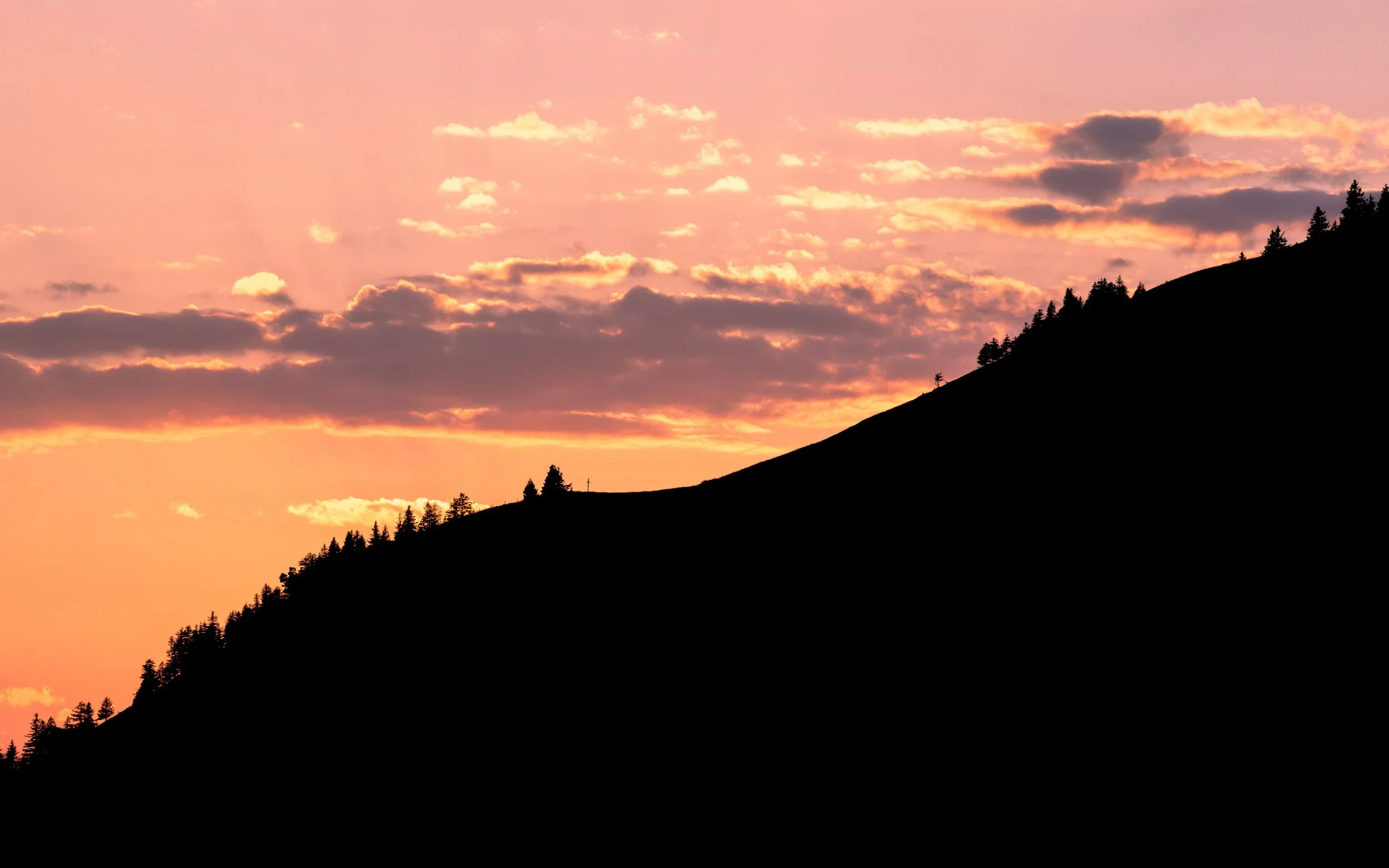 Sunset over a mountain slope with silhouettes of trees and a few clouds in pink, orange, and purple sky.