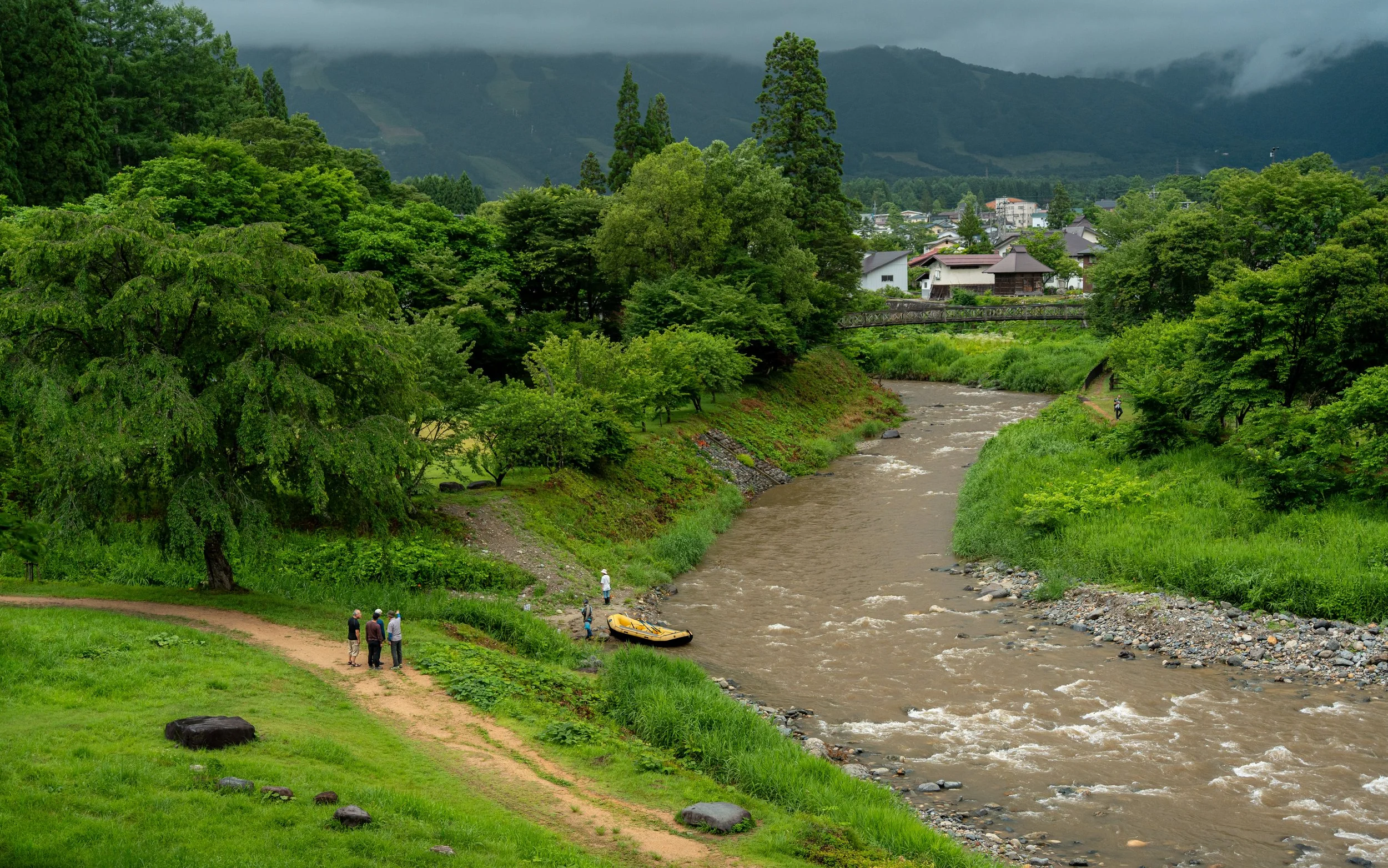 A lush green park with a walking path and a small group of people near the riverbank, a boat is on the shore, with houses and trees in the background and mountains under a cloudy sky.