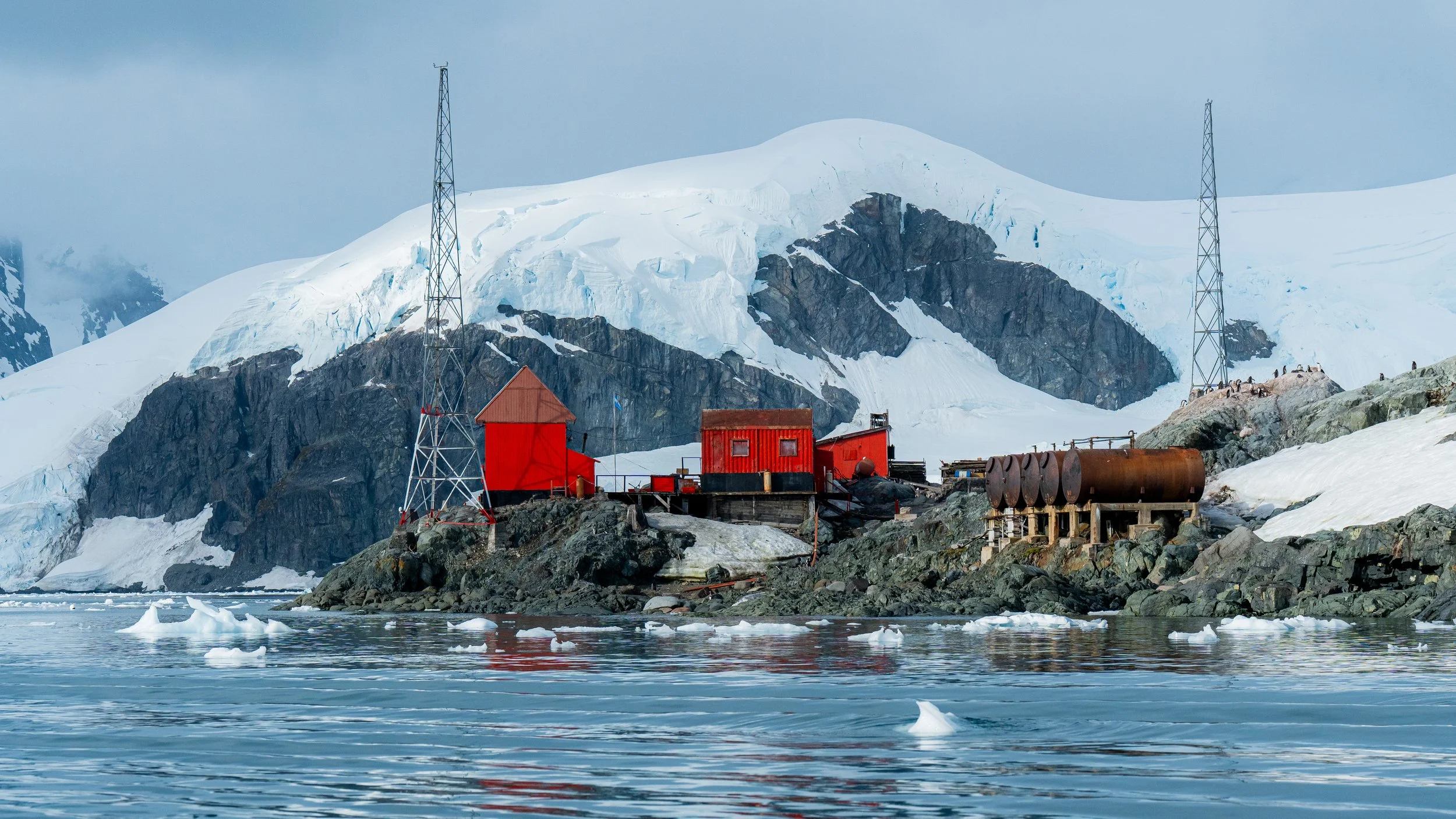 A small research station with red buildings on a rocky shoreline by icy water, set against a snowy mountain with glaciers and ice. Two communication towers are on either side of the station, and penguins are visible on the rocks.