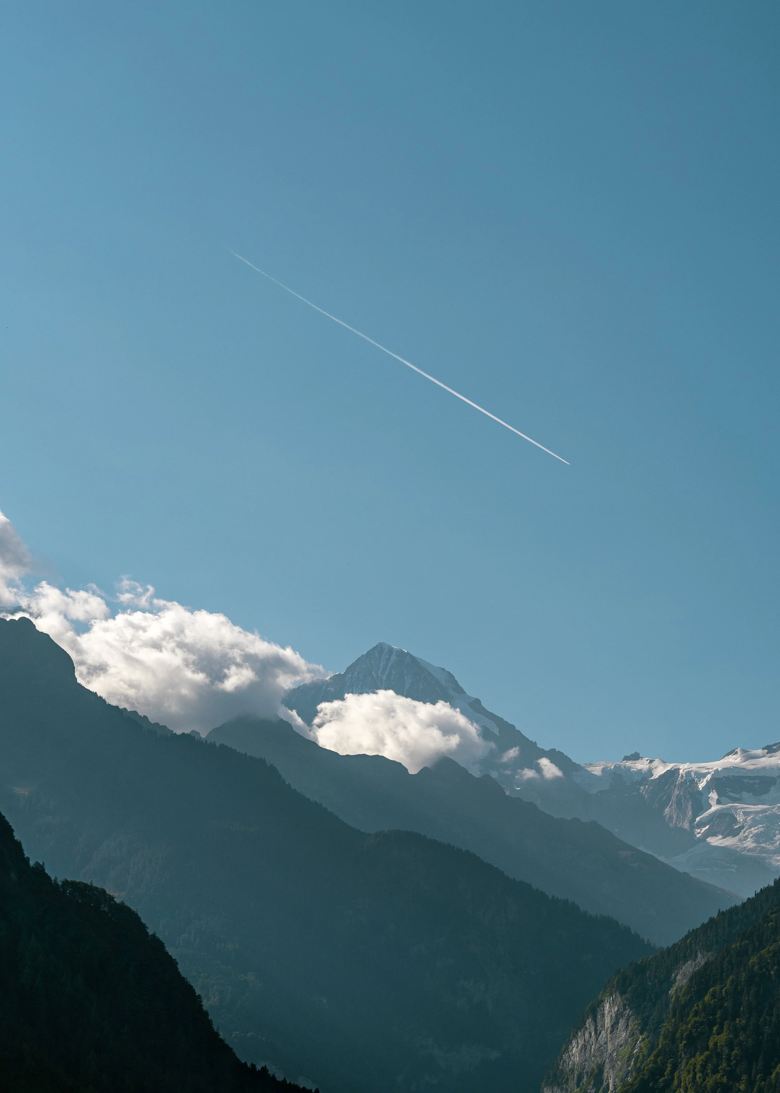 Snow-capped mountain peaks in the background with partly cloudy sky, green forested mountain slopes in the foreground, and a contrail in the blue sky.