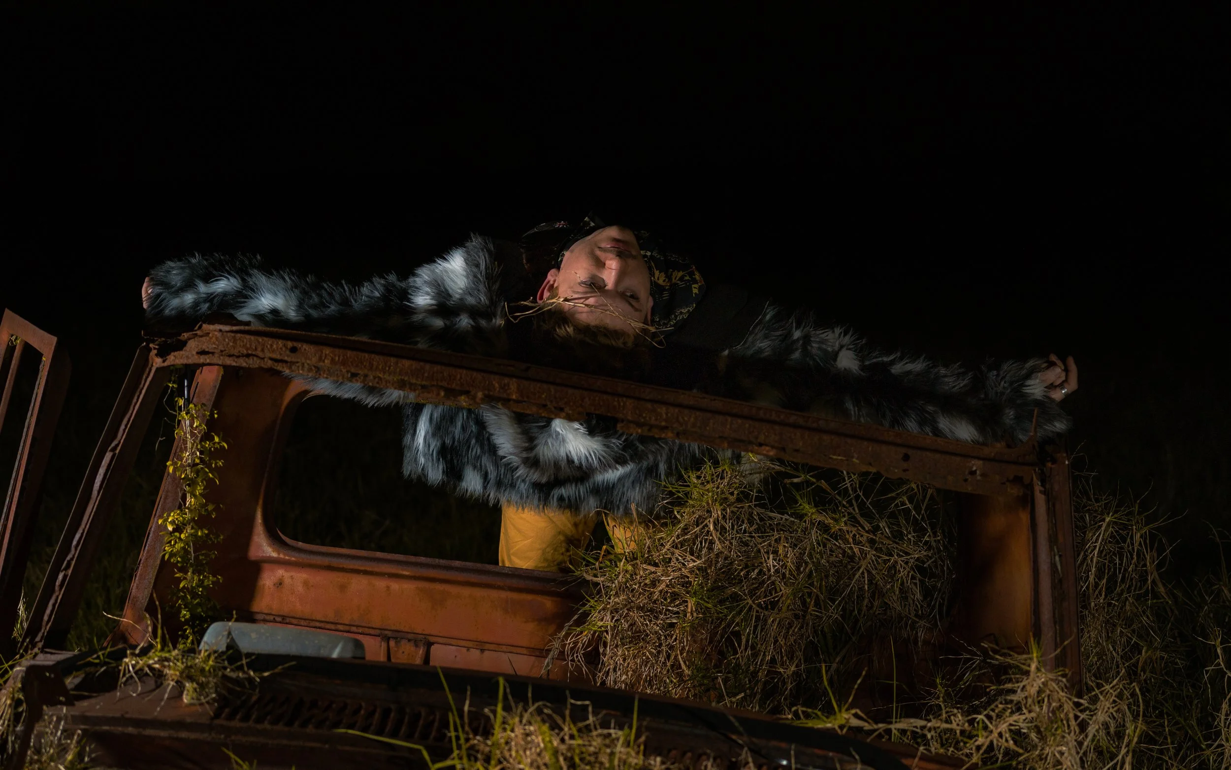 A person lying on top of an old, rusty car with grass and plants growing around it, night scene with dark sky.