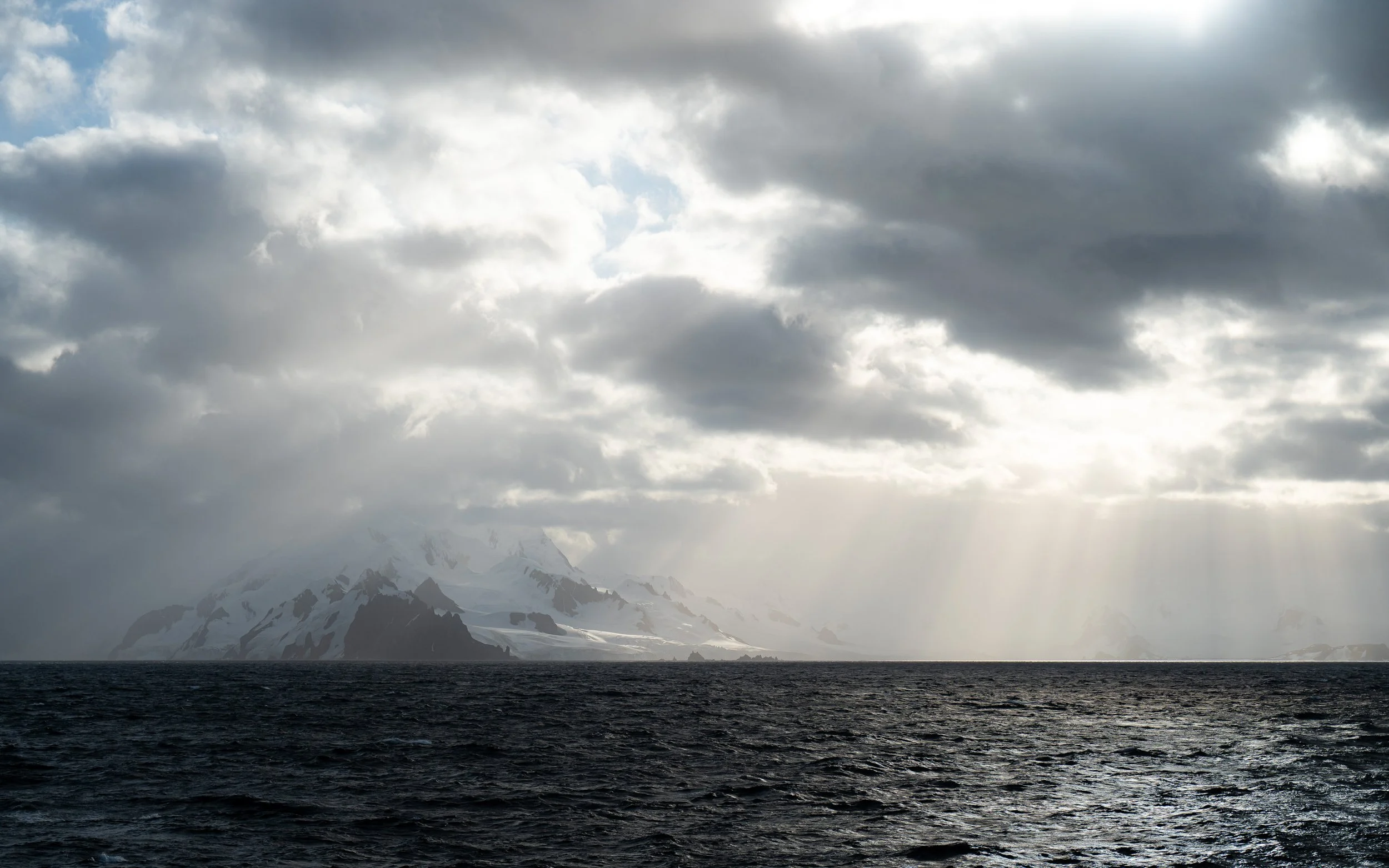 A vast ocean under a cloudy sky with mountain peaks visible in the background, partly obscured by mist or fog.
