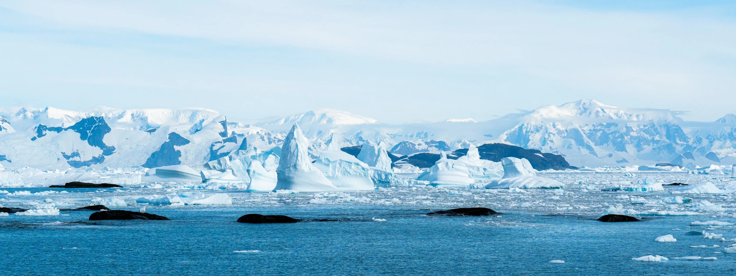 A glacial landscape with icebergs floating on the ocean and snow-covered mountains in the background.