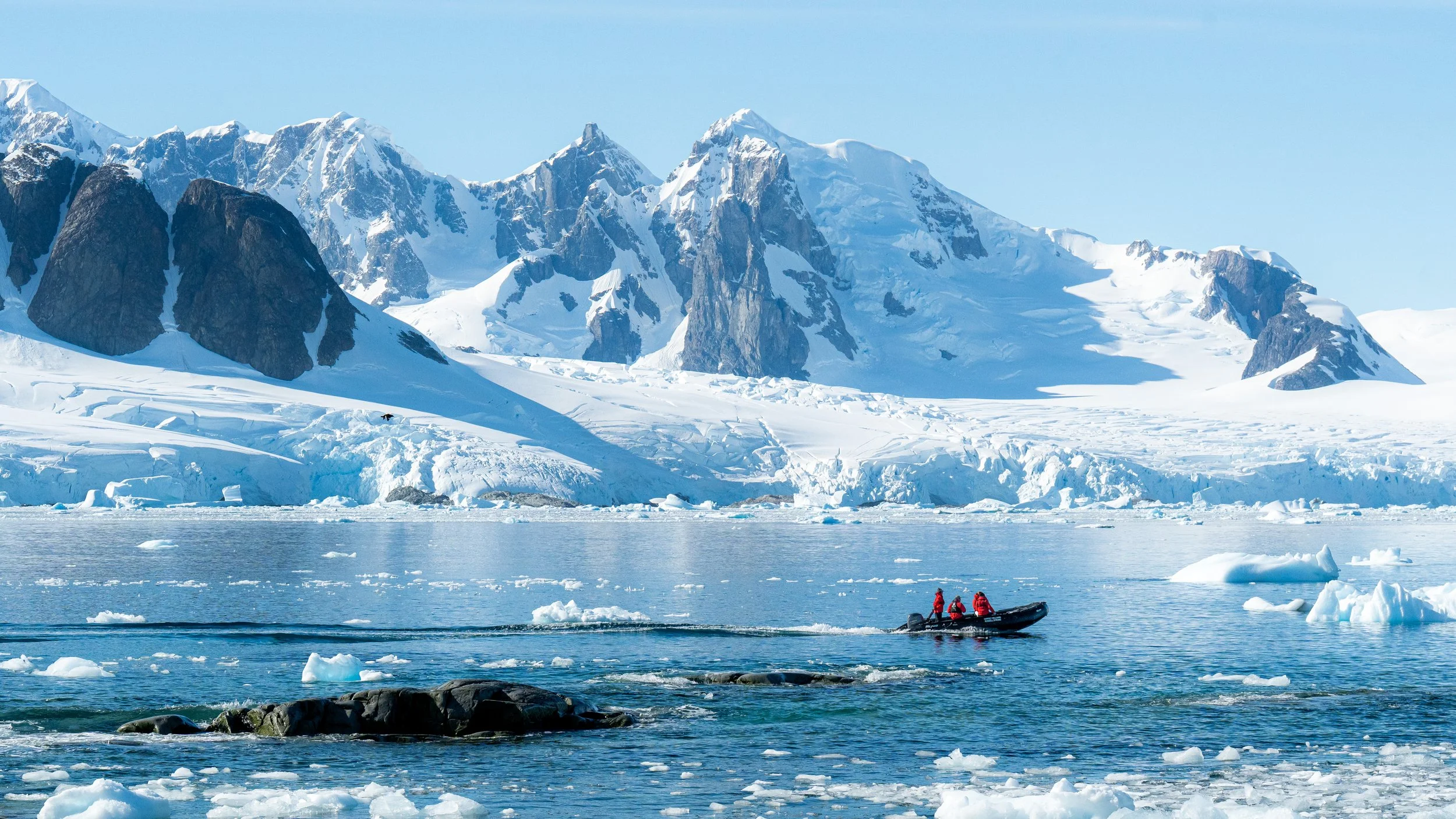 A boat with four people dressed in red jackets navigates through icy water near glacial mountains covered in snow.