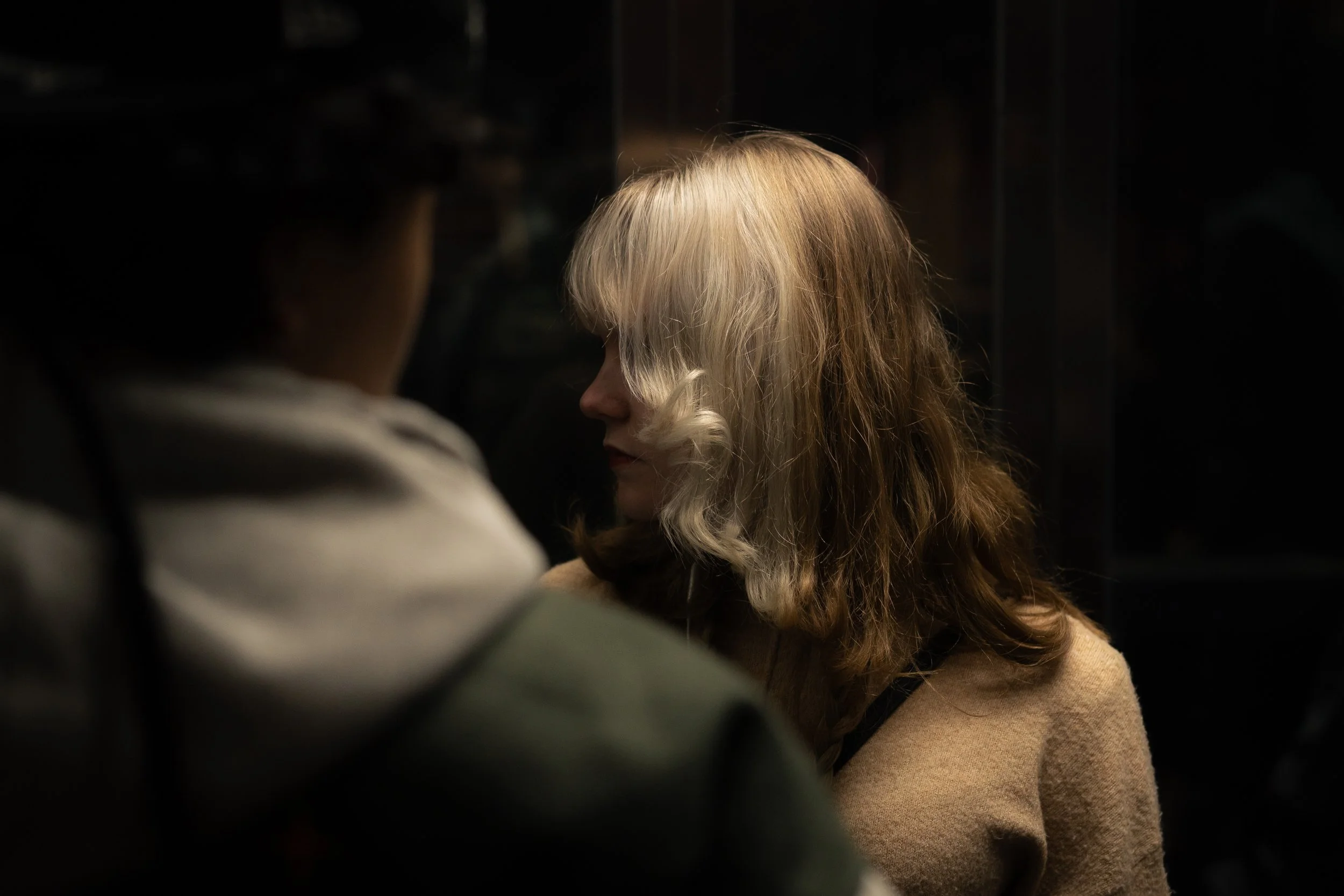 Side view of a blonde woman with wavy hair wearing a tan sweater, sitting in a dimly lit environment, with another person's back visible in the foreground.