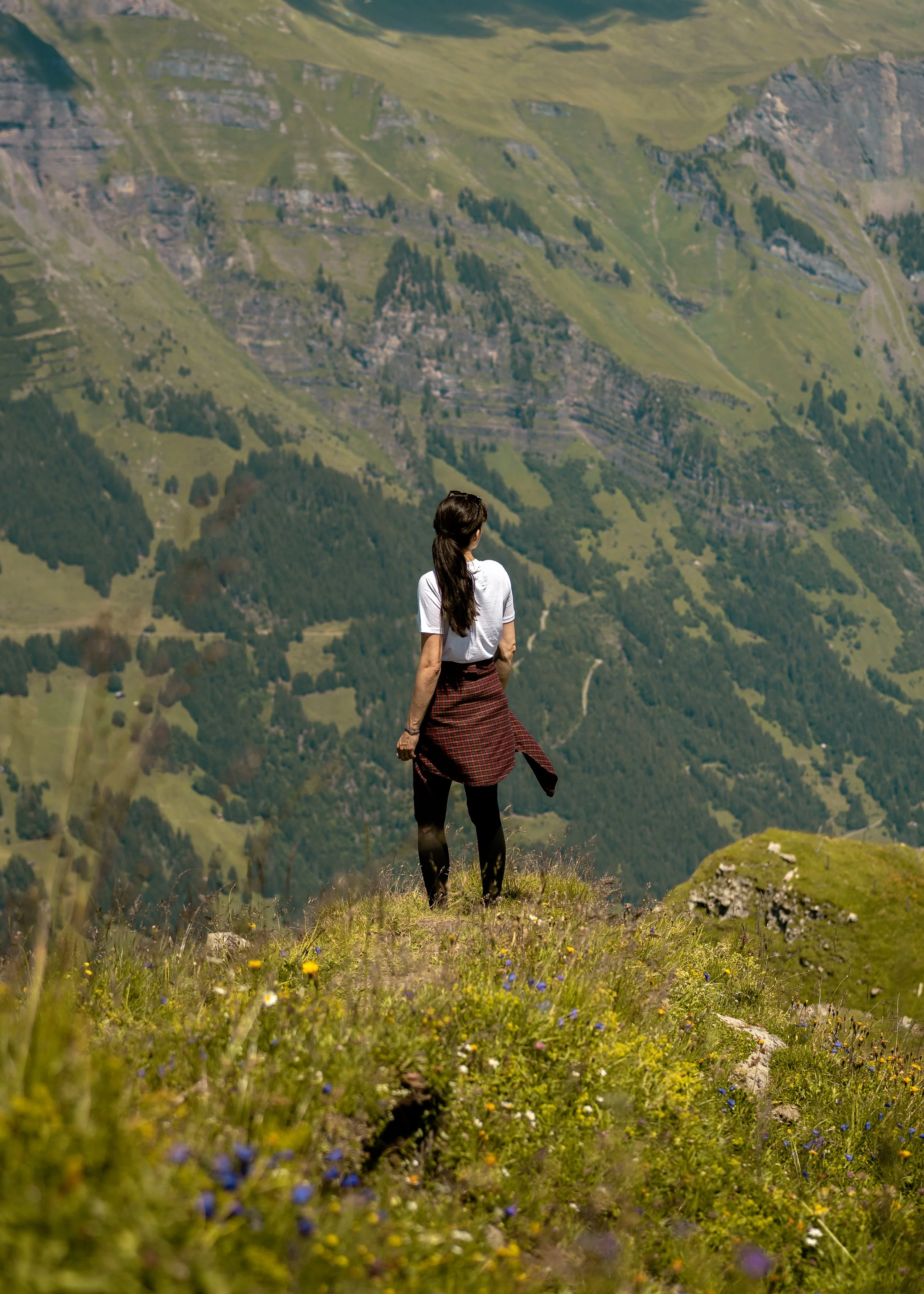 A woman with long brown hair, wearing a white shirt and patterned skirt, standing on a grassy hillside overlooking lush green mountains with terraced fields and patches of trees.