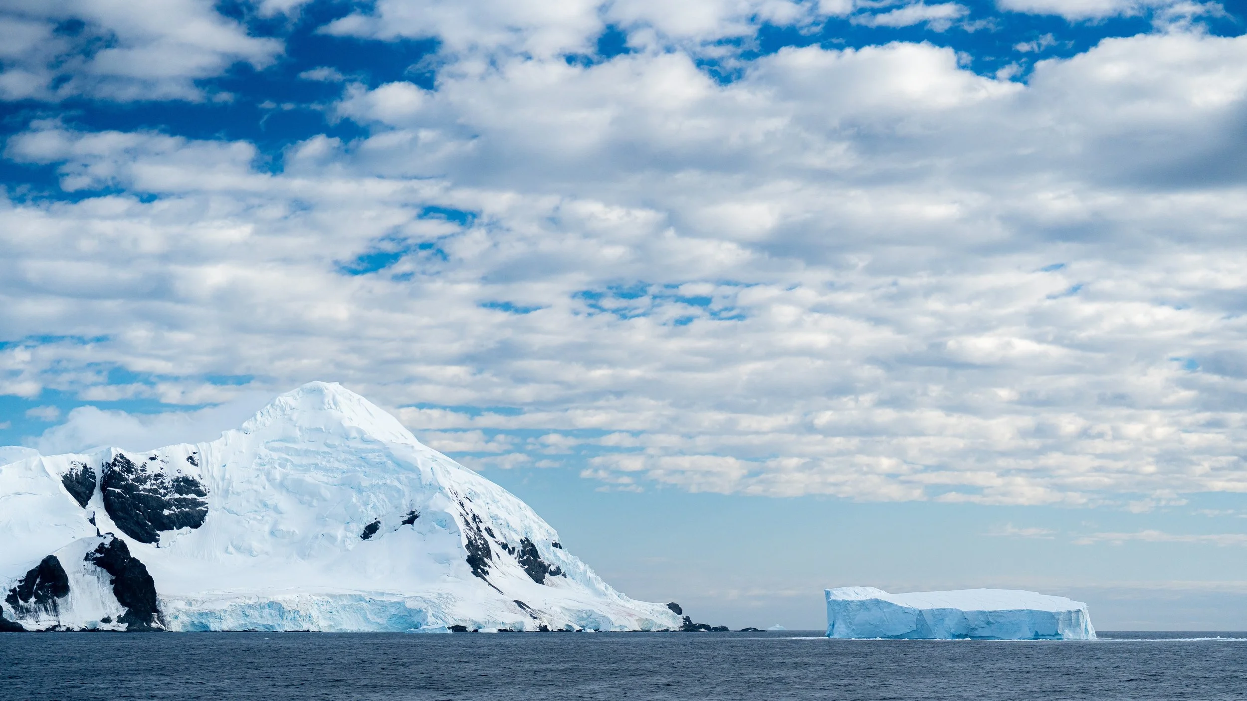 Snow-covered mountain with ice formations in the ocean and a partly cloudy sky.