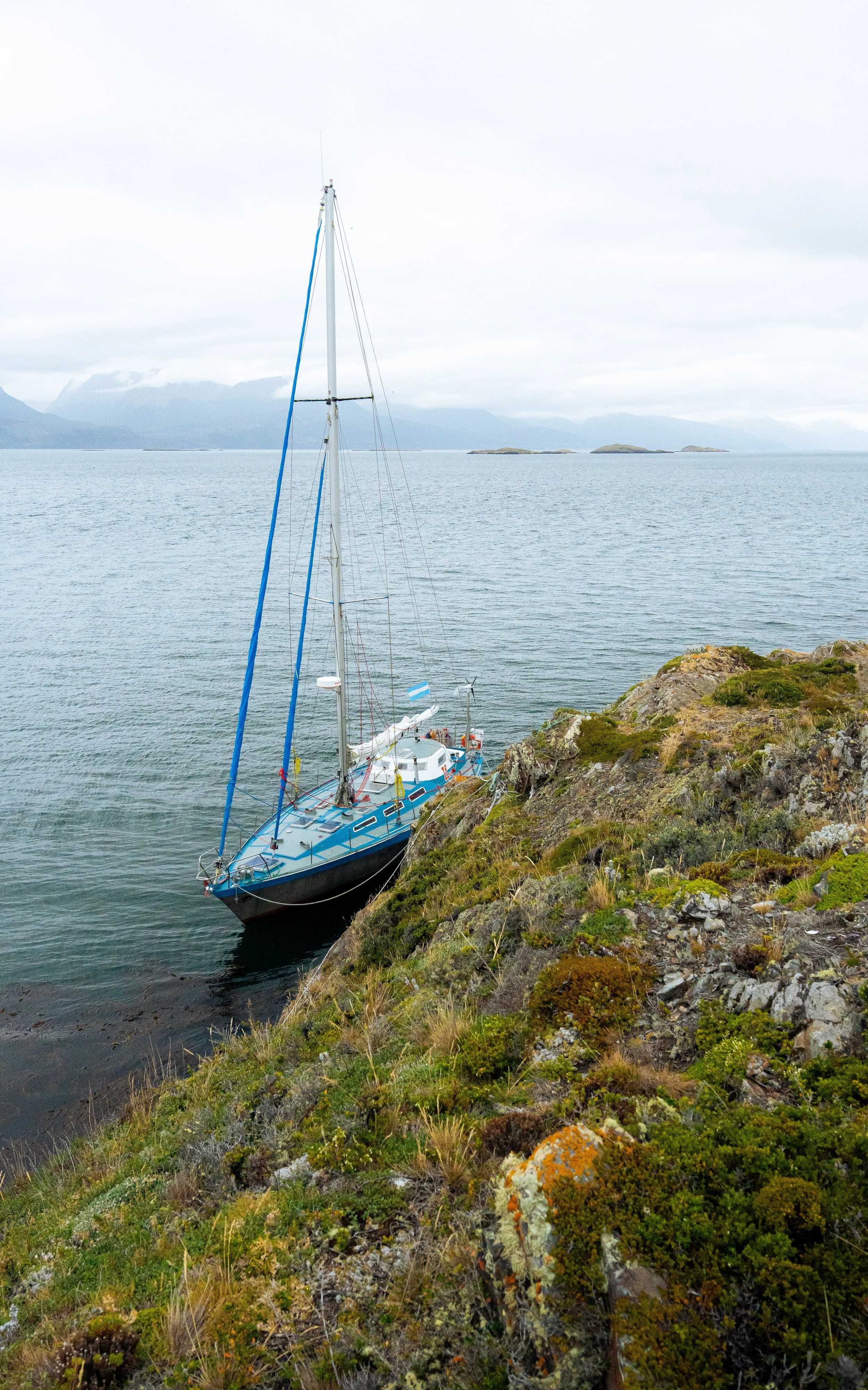 A sailboat docked near a rocky and mossy shoreline with distant islands and mountains under a cloudy sky.
