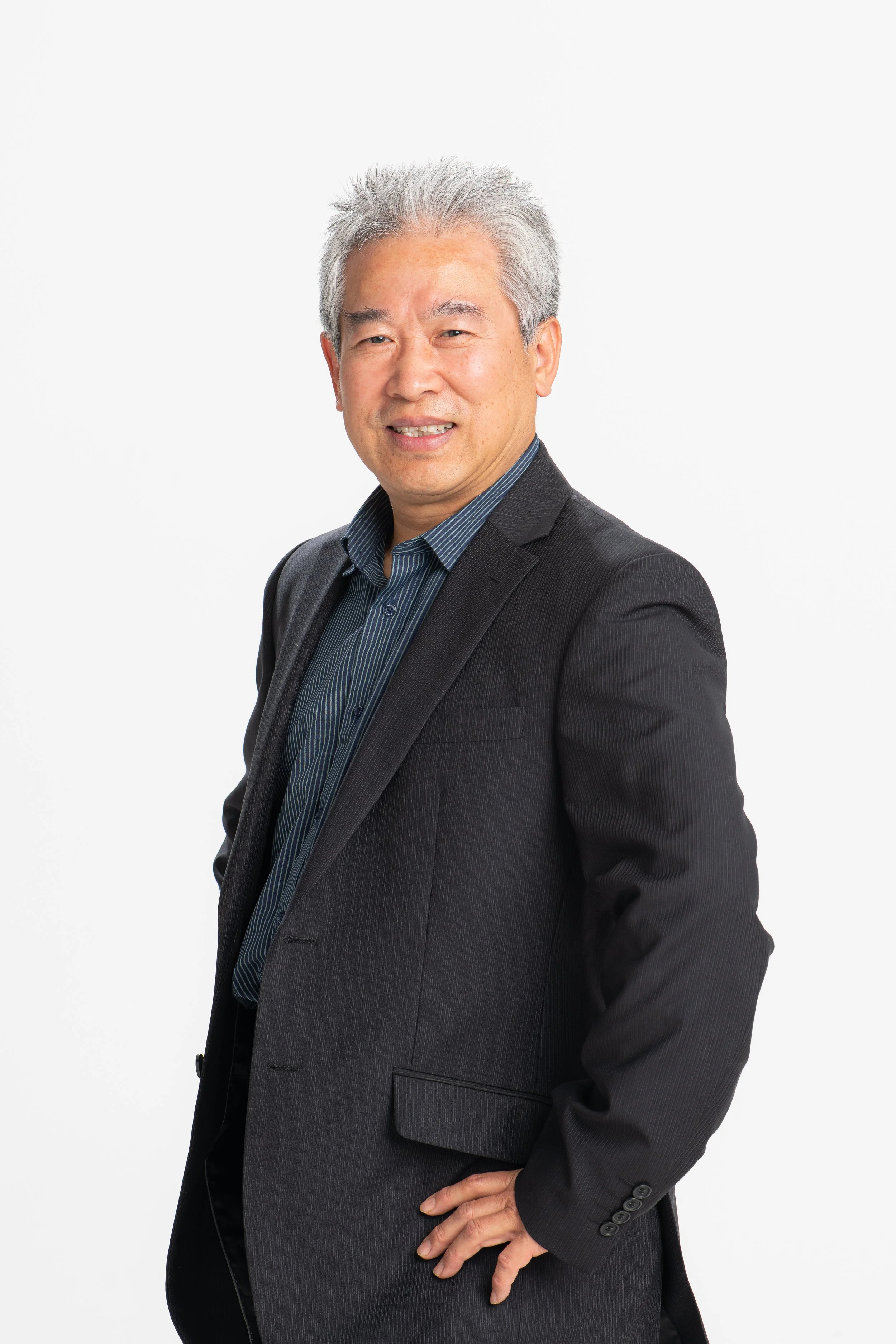 Portrait of a middle-aged man with gray hair wearing a black suit jacket over a blue striped shirt, smiling and posing with one hand on his hip against a plain white background.