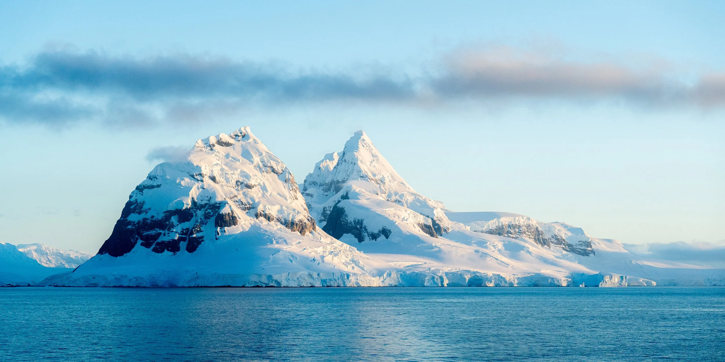 Snow-covered mountains with jagged peaks rising above icy waters under a partly cloudy sky.