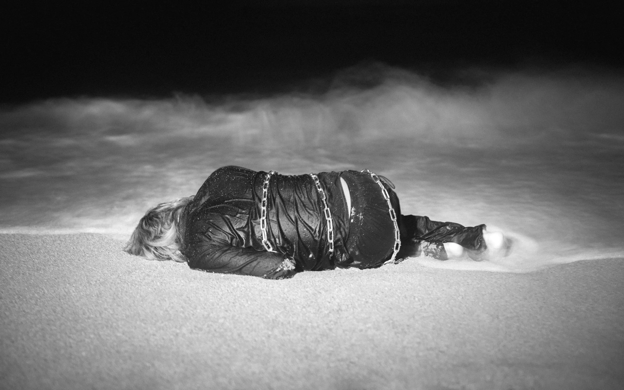 A person lying face down on a sandy beach at night, wearing a wet jacket and chain, with the ocean waves in the background.