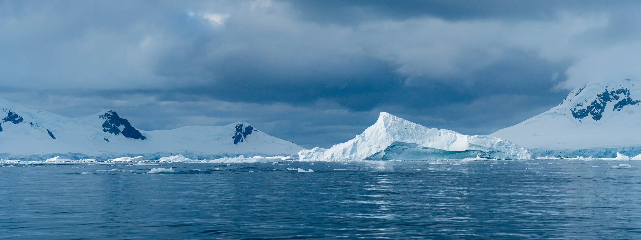 Aerial view of icy Antarctic landscape with snow-covered mountains, floating icebergs, and a large iceberg in the water under a cloudy sky.